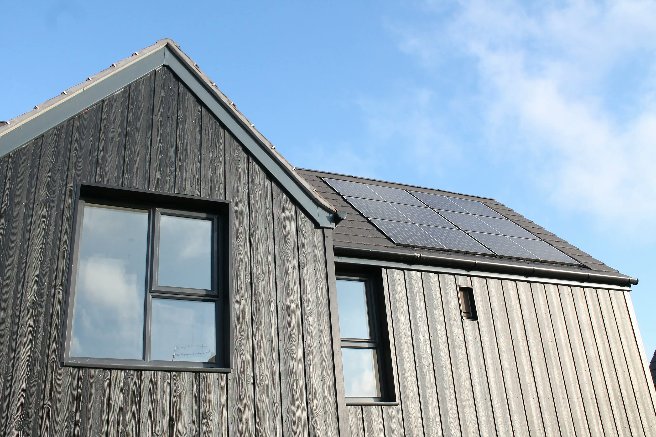 A contemporary single-storey house with a dark timber façade and expansive floor-to-ceiling windows, set beneath a clear blue sky with scattered clouds. The slanted roof is fitted with sleek solar panels, emphasising sustainability.