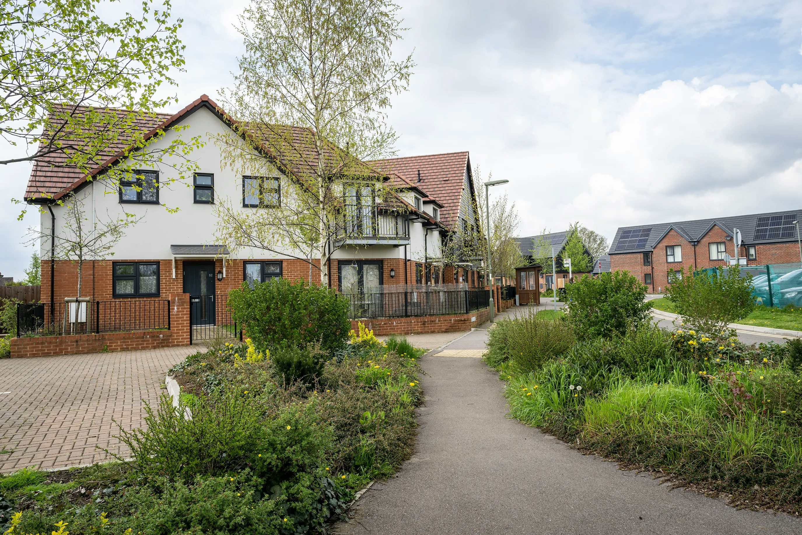 A modern residential street with semi-detached houses featuring a mix of red brick and white-rendered facades. The nearest house has a red-tiled, pitched roof and a paved driveway bordered by a neat garden with flowering shrubs and ornamental grasses. Further houses line the background, some with solar panels on the roofs, under a partly cloudy sky that casts soft, natural light over the well-kept neighbourhood. The scene suggests a quiet, eco-conscious community.
