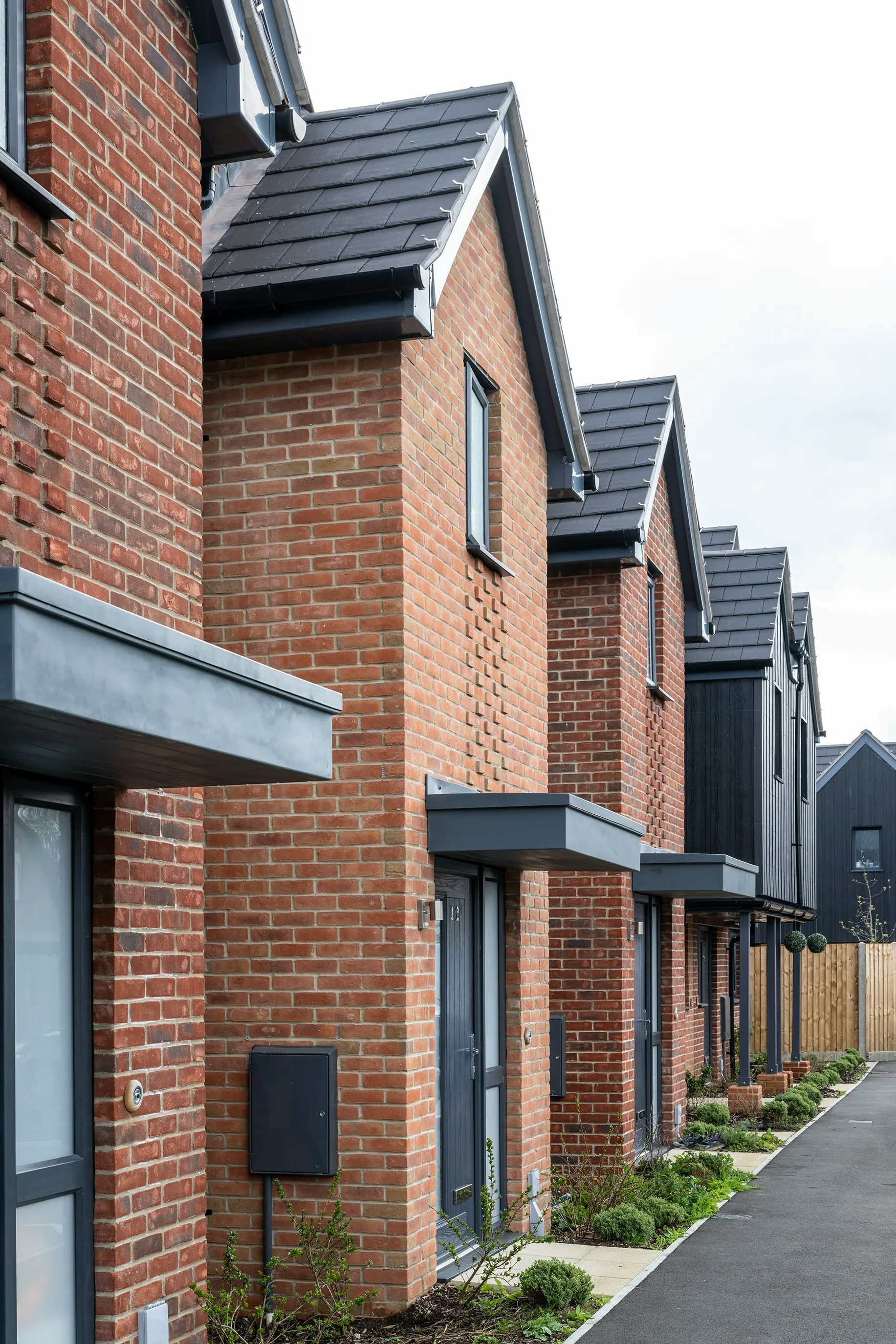 Row of contemporary two-storey brick houses with dark grey tiled roofs and black-framed windows, each with small landscaped front gardens and paved paths leading to their entrances, creating a clean, uniform residential streetscape.