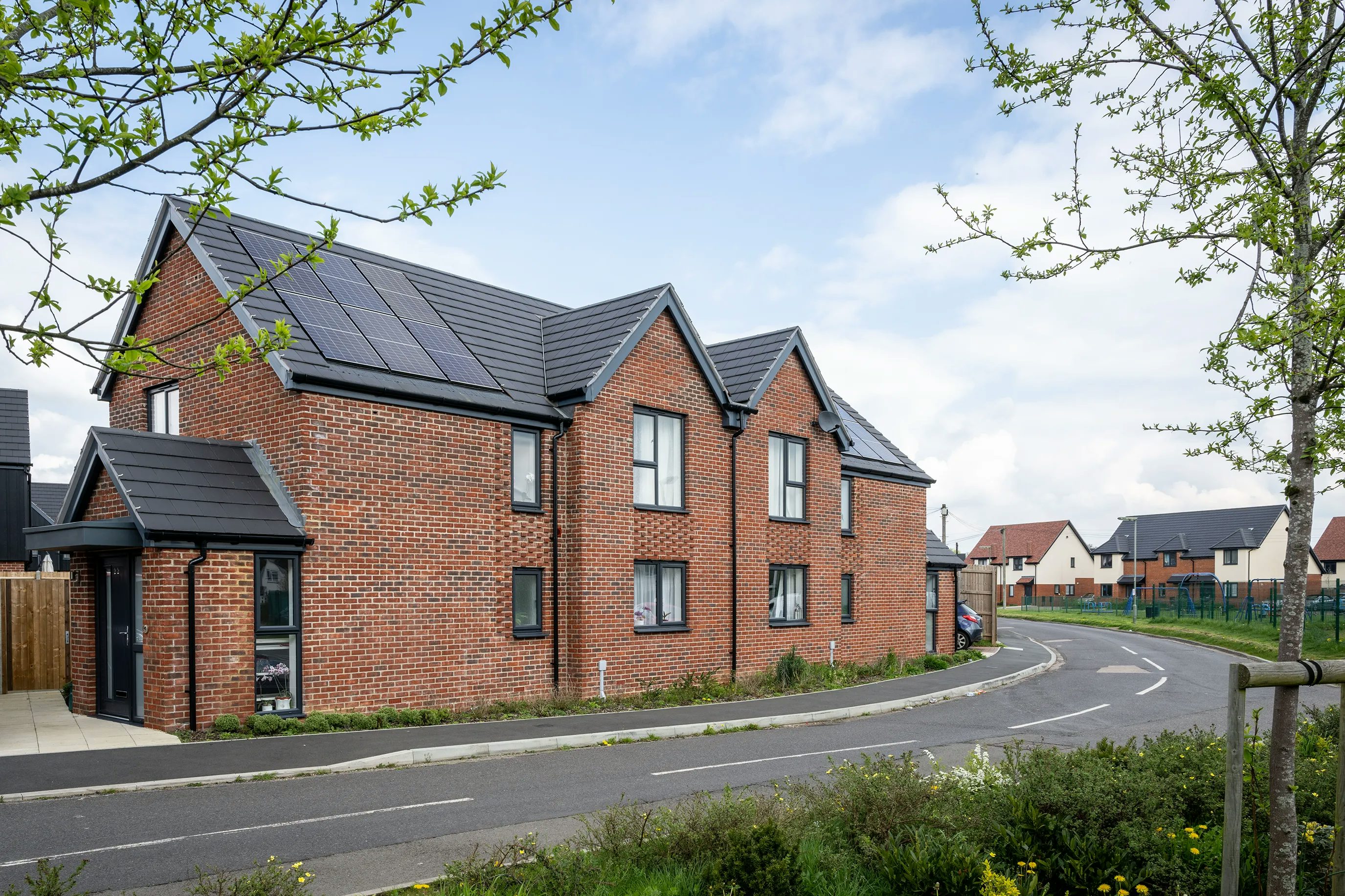 Curved residential street lined with newly built brick houses, each featuring dark grey roofs fitted with solar panels. The scene is framed by neat greenery—trees, bushes, and well-kept lawns—under a partly cloudy sky, evoking a clean, sustainable, and modern neighbourhood atmosphere.