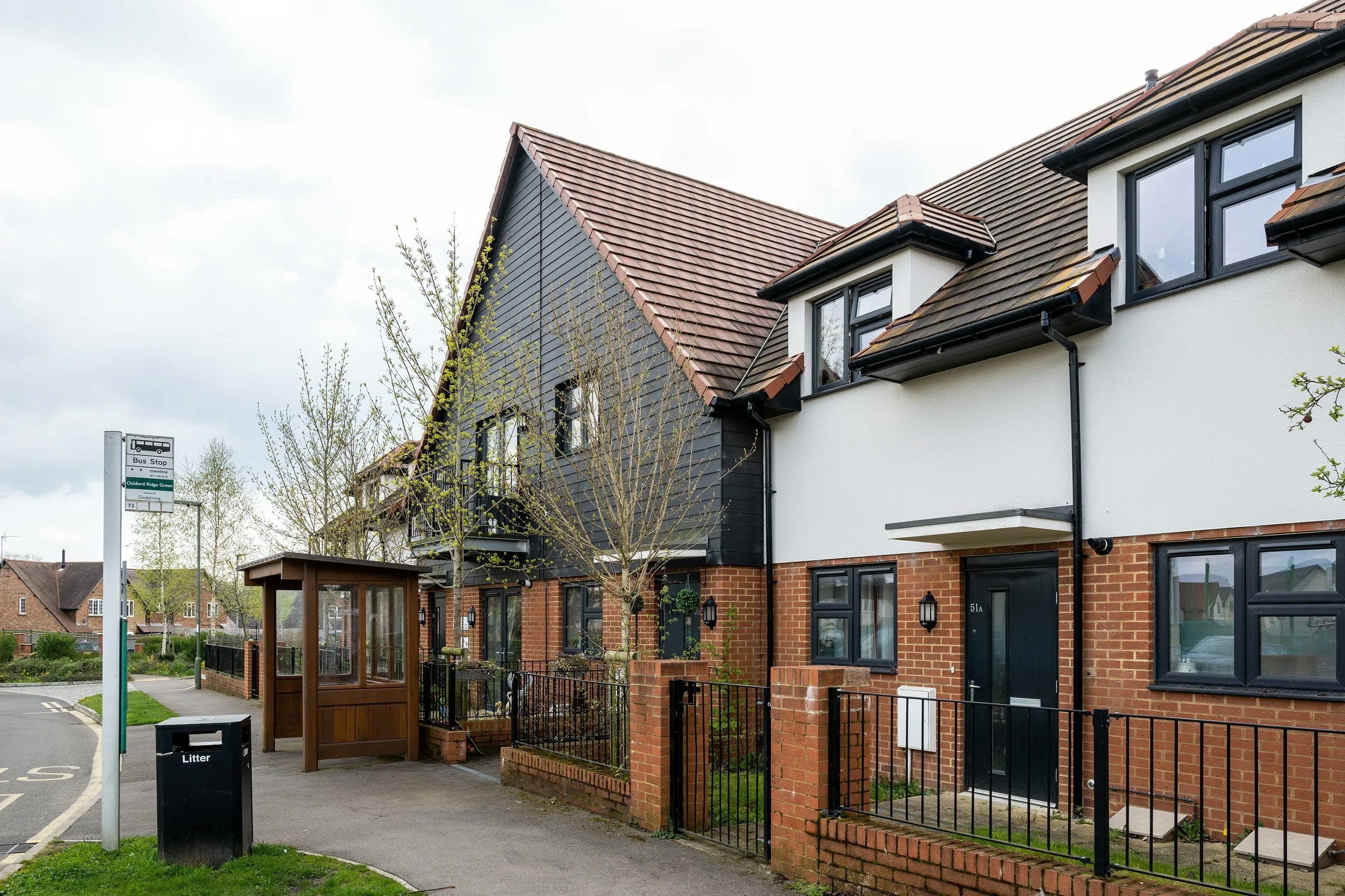 A quiet residential street with modern townhouses featuring white and brick façades and dark roofs. A covered bus stop with a litter bin and a sign for route 2 stands on the pavement. The area is clean and green, with trees in the background and no people present.