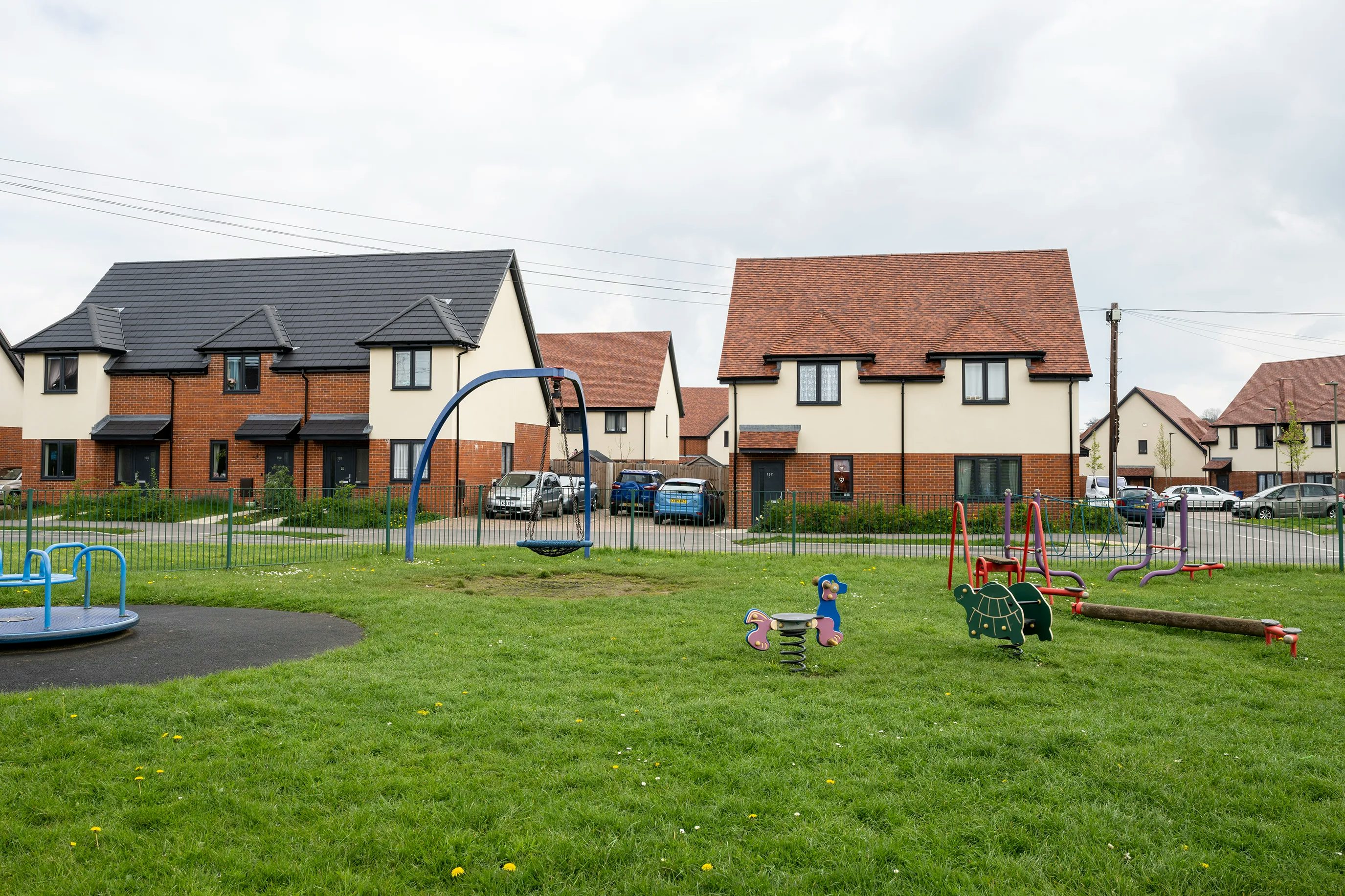 A community playground nestled within a modern residential development on a cloudy day. The scene features a green lawn with scattered yellow wildflowers, surrounding play equipment including a merry-go-round, swings, and animal-shaped spring riders. Contemporary houses with brick and cream façades line the background, with several cars parked along the curb, creating a calm and family-friendly atmosphere.