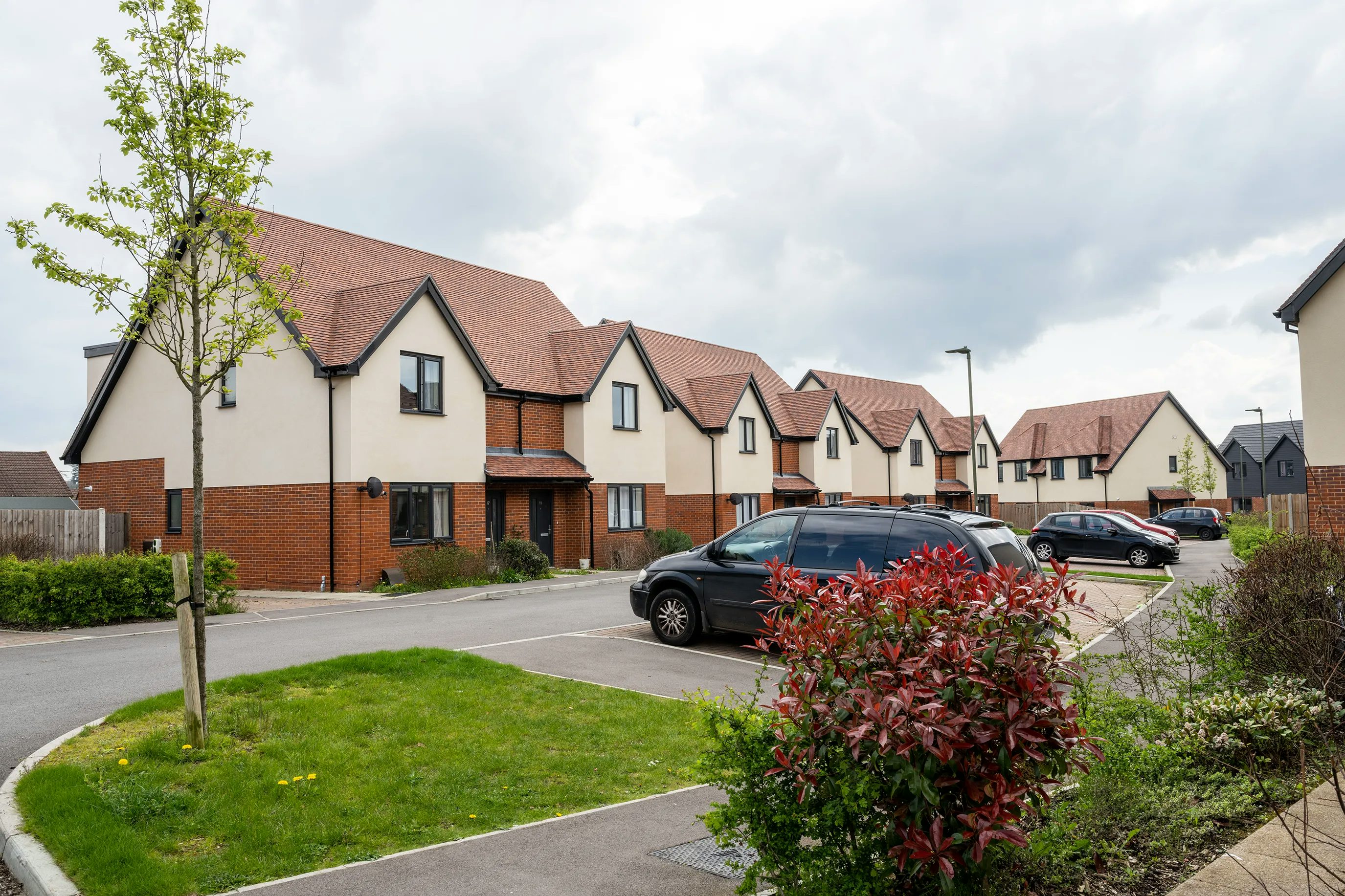 A residential street lined with modern suburban houses featuring pitched roofs and façades of brick and cream render. Several cars are parked in designated spaces outside the homes, while neat grass verges and small trees provide a landscaped setting. The overcast sky casts a subdued light, contributing to a quiet, everyday atmosphere. The scene reflects contemporary housing design and orderly suburban living.