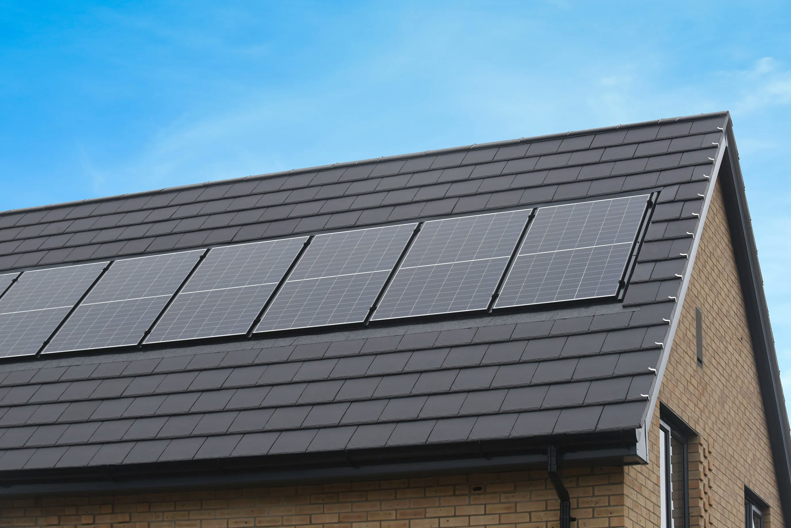Modern house with six rooftop solar panels arranged in two rows on a dark grey tiled roof, under a clear blue sky on a sunny day.