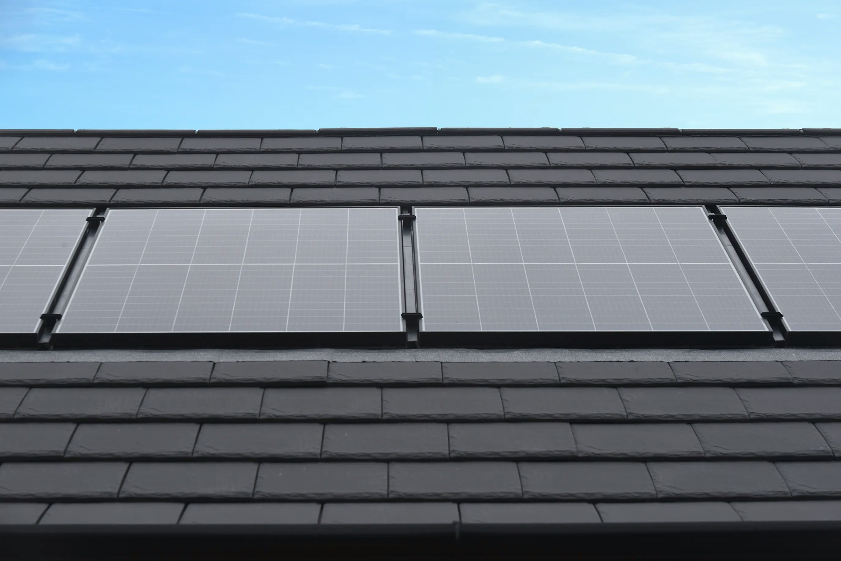 A row of dark, rectangular solar panels is mounted on a sloped rooftop covered with asphalt shingles. The panels are aligned neatly under a clear blue sky, reflecting subtle sunlight. The scene conveys a focus on renewable energy and sustainable residential design, with no visible background distractions.