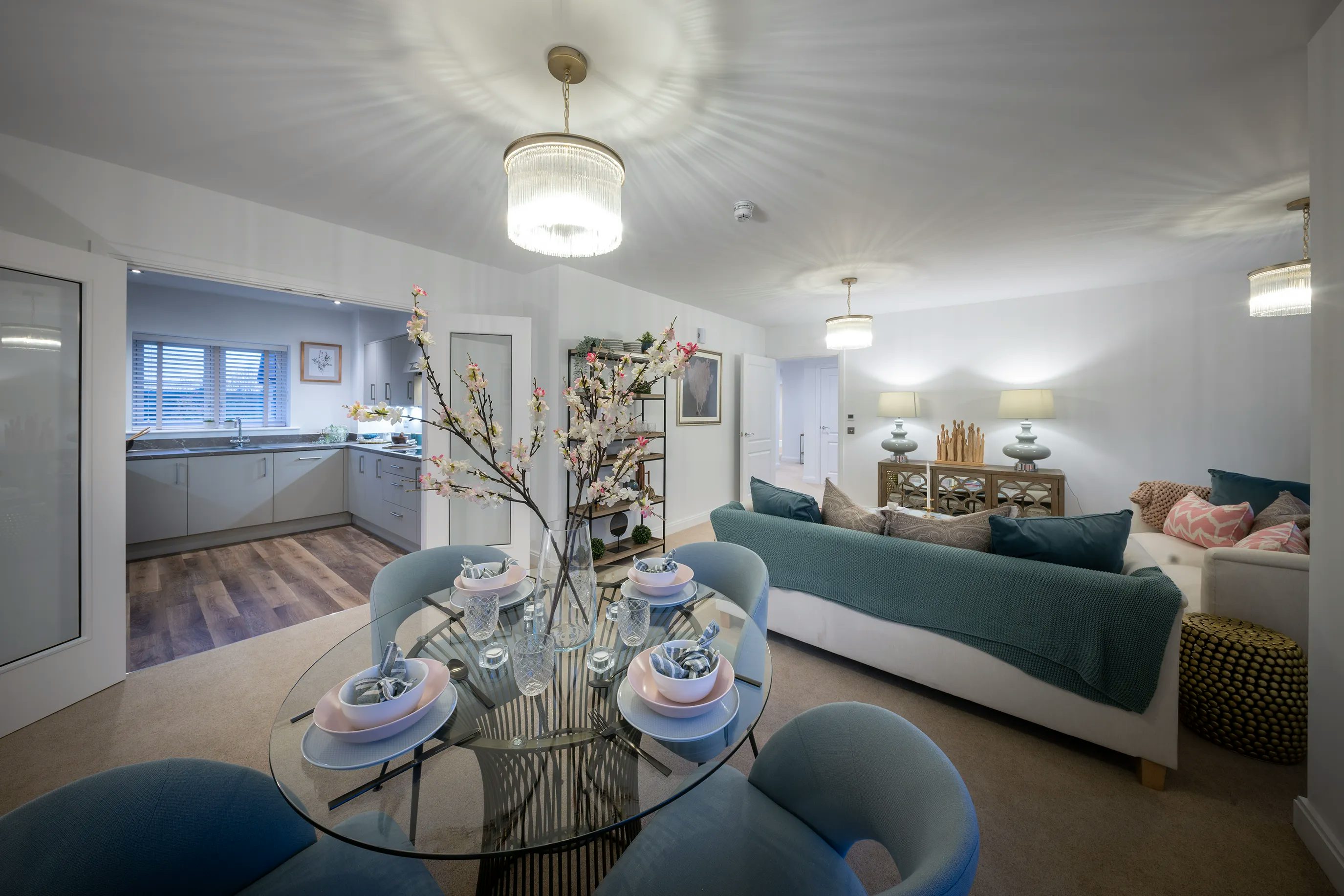 A modern, open-plan living and dining space featuring a round glass dining table set for four, with elegant tableware and a floral centerpiece. The adjoining living area includes a large sofa with blue and pink cushions, warm ambient lighting, and a side table with lamps and decorative accents. In the background, a contemporary kitchen is partially visible, adding to the cohesive and inviting atmosphere of the room.