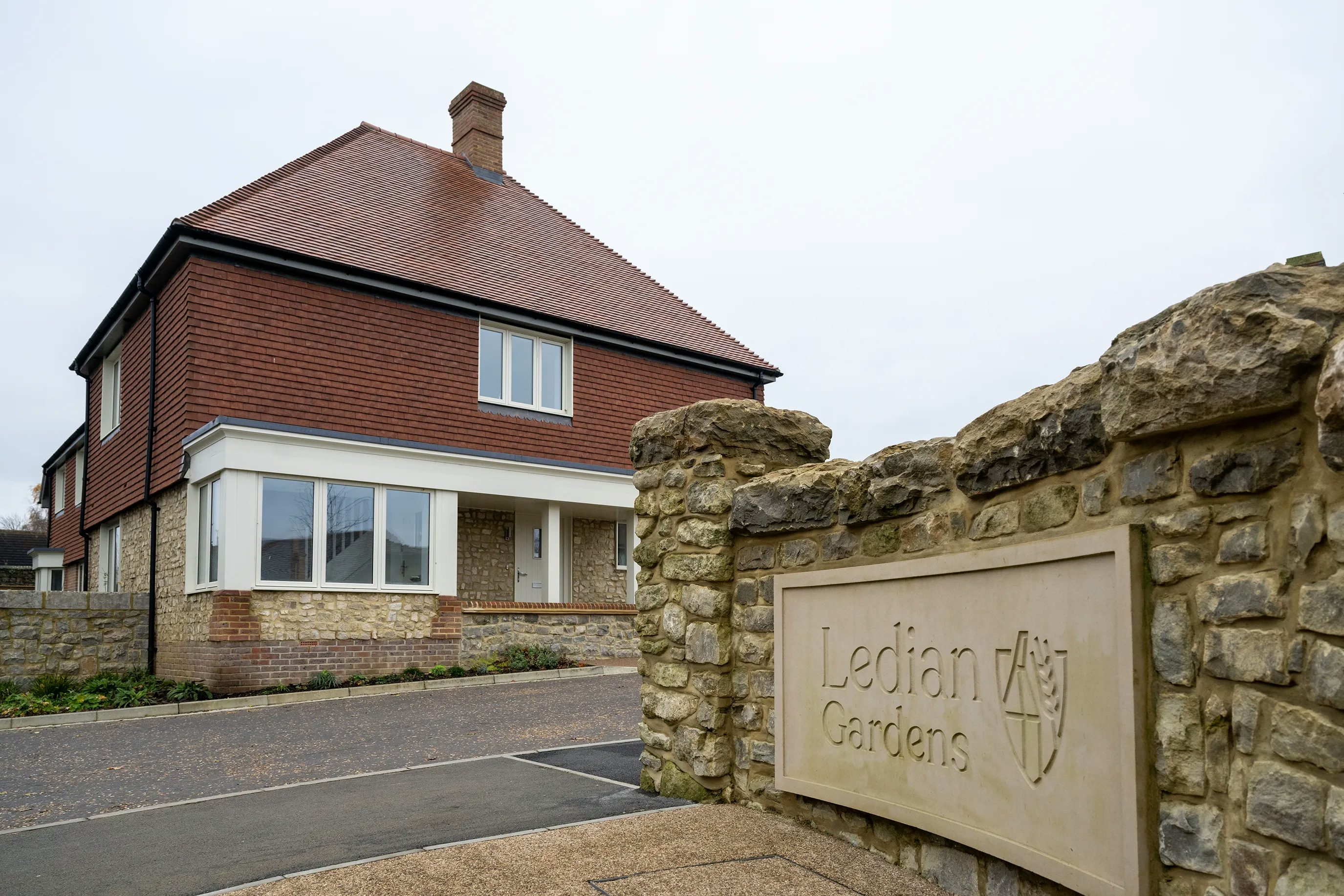 A two-story residential house with a red-tiled roof and a mixed brick and stone facade. A bay window features prominently on the ground floor, and a chimney rises from the roof. In front of the house, a stone wall bears a shield emblem and the text “Ledian Gardens,” indicating the property’s name or development.