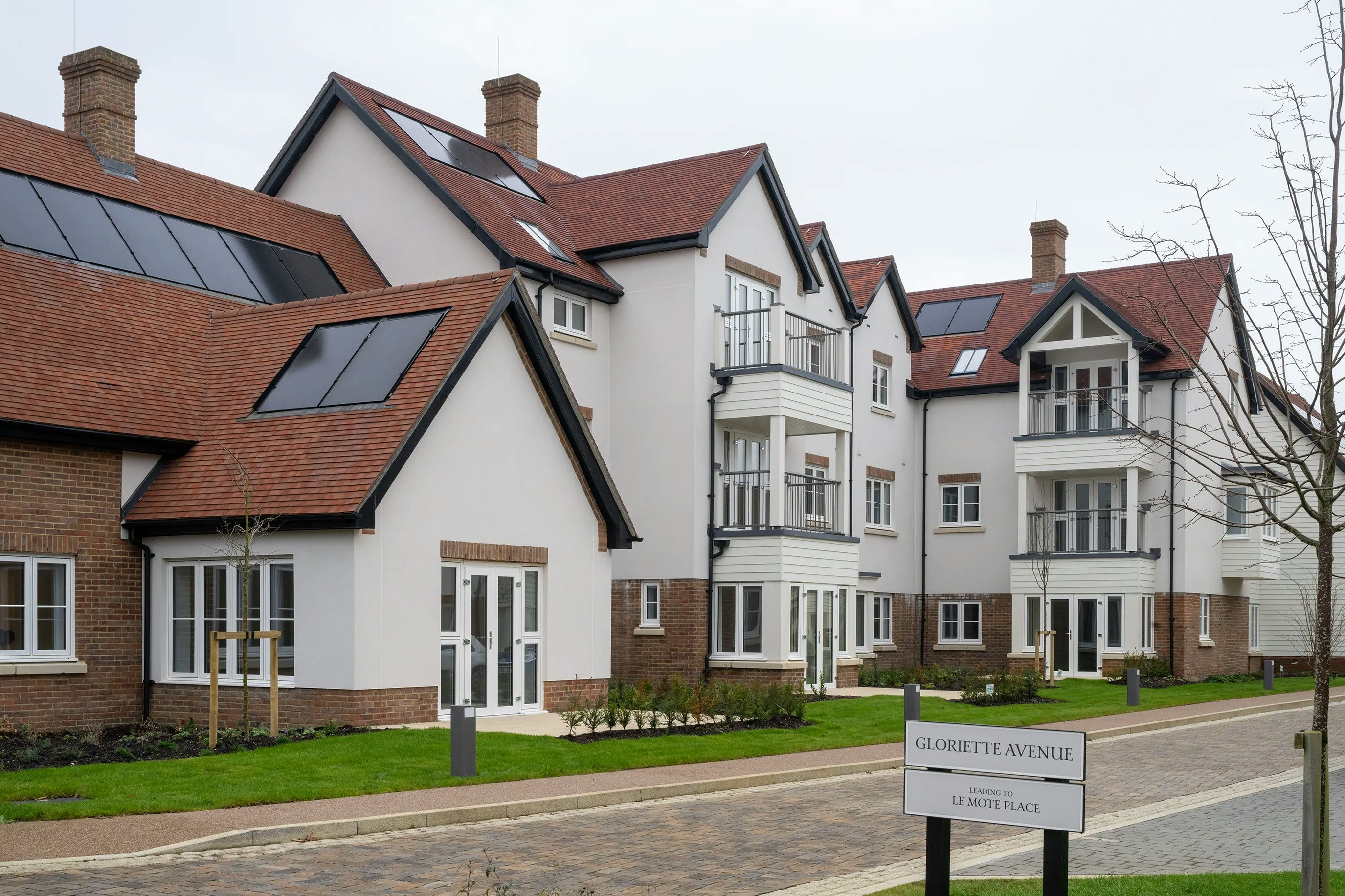 A modern residential complex featuring white façades with warm brick accents and red-tiled roofs. Rooftop solar panels suggest a focus on sustainability. The buildings include balconies, expansive windows, and are surrounded by neatly maintained green spaces with small trees. In the foreground, a sign reads “GLORIETTE AVENUE” and “LE MOTE PLACE,” indicating the development’s location and name. The overall atmosphere is clean, contemporary, and community-oriented.