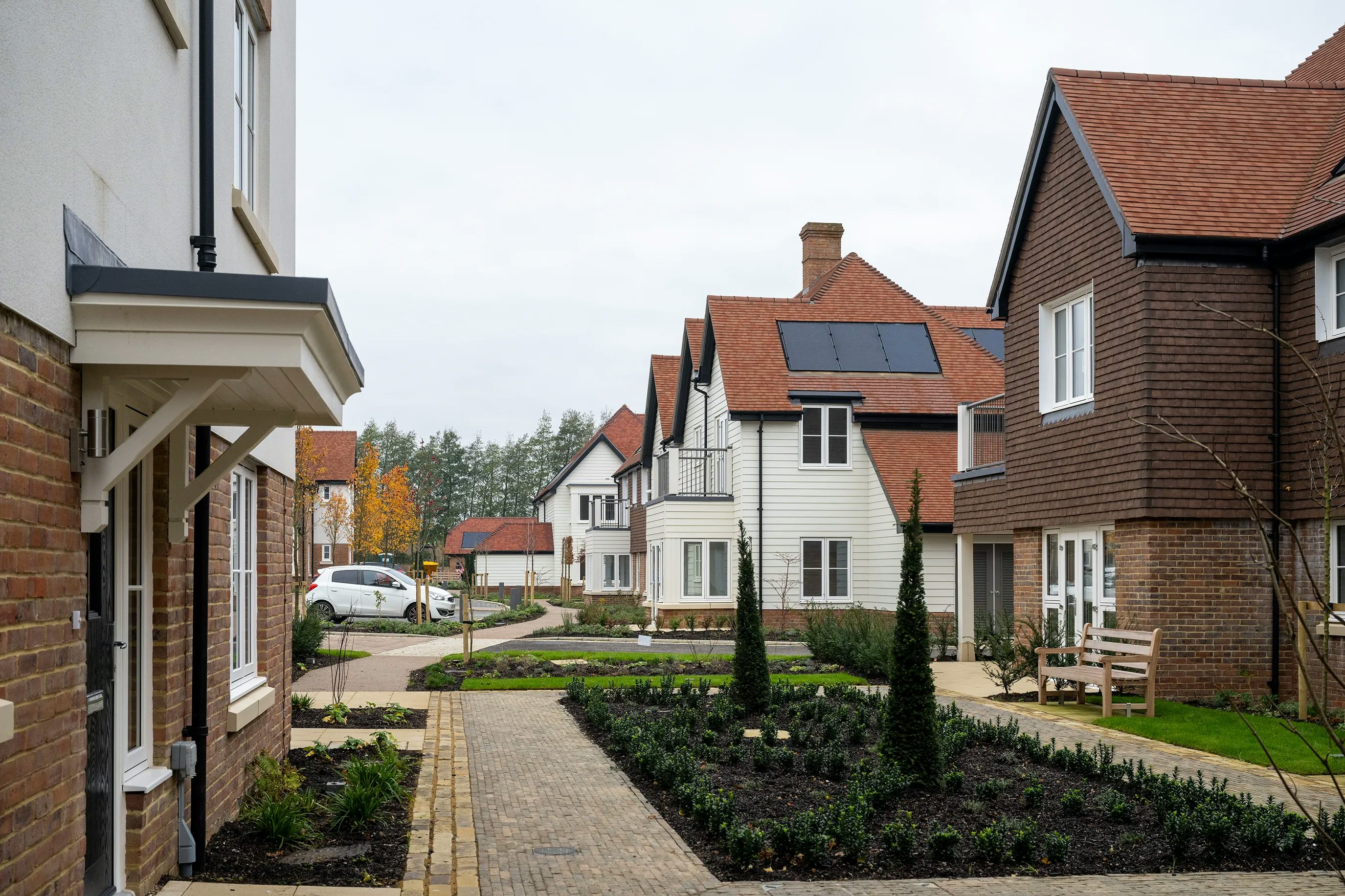 Modern residential street with brick and white-sided houses, some featuring rooftop solar panels. A white car is parked on the roadside, and the foreground includes a landscaped path bordered by small trees and neatly trimmed bushes, evoking a calm and sustainable suburban atmosphere.