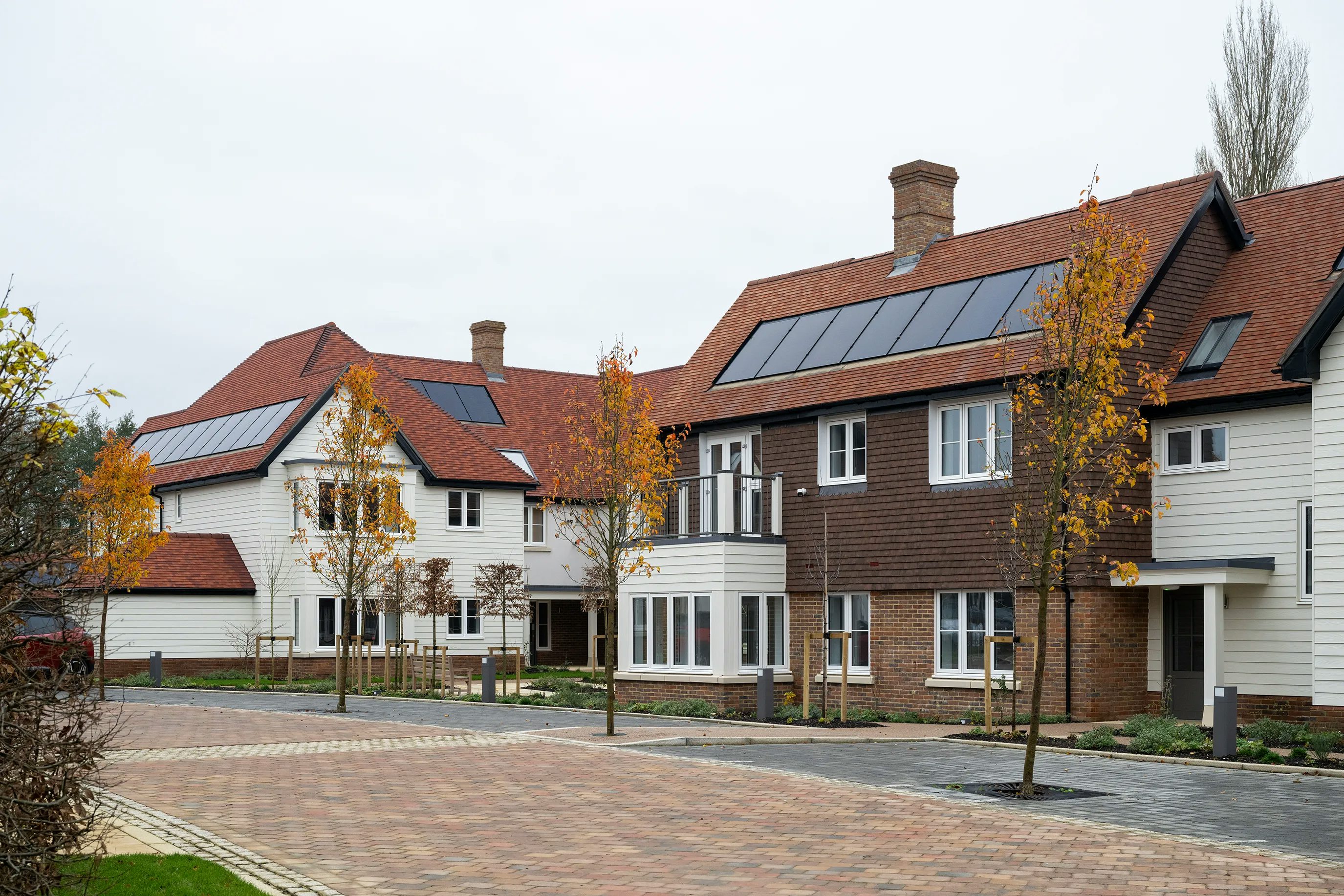 A row of modern, semi-detached houses with red-tiled roofs and solar panels, lining a clean, paved residential street. The façades feature a mix of white and brown finishes, large front-facing windows, and planted young trees along the pavement, evoking a sense of sustainable, community-focused living.