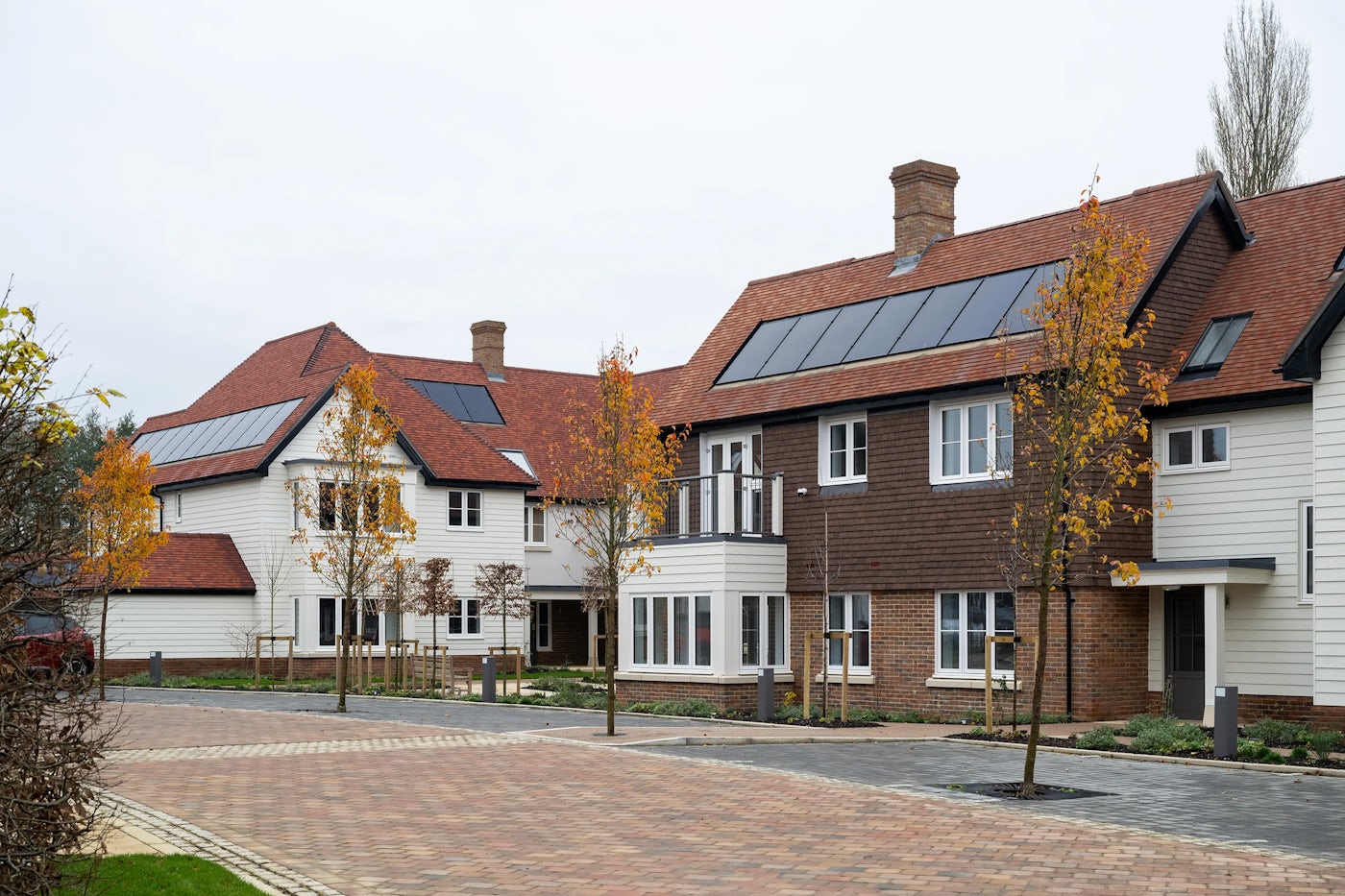 A row of modern, semi-detached houses with red-tiled roofs and solar panels, lining a clean, paved residential street. The façades feature a mix of white and brown finishes, large front-facing windows, and planted young trees along the pavement, evoking a sense of sustainable, community-focused living.