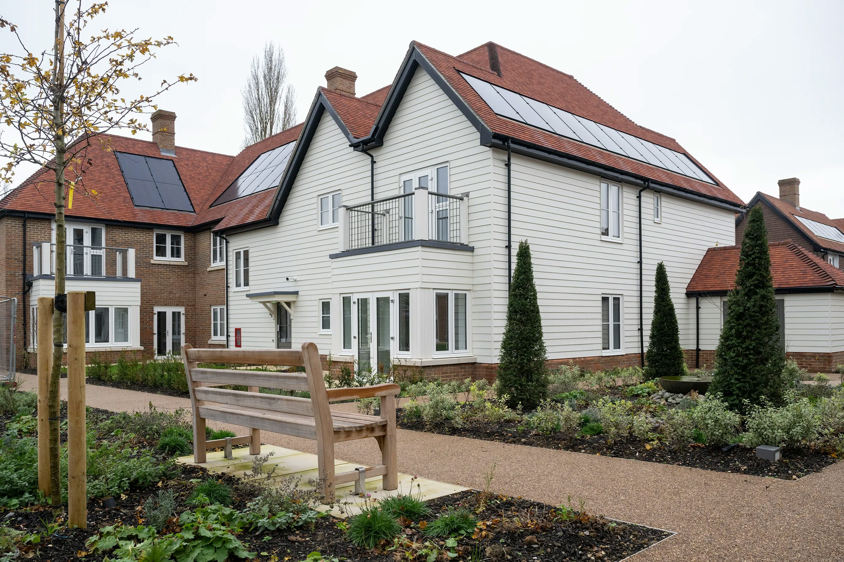Modern residential building with white cladding, red-tiled roof, and rooftop solar panels. In the foreground, a small landscaped garden features varied plants and a wooden bench, suggesting a communal or private outdoor space. The building includes multiple windows and a balcony, hinting at comfortable, energy-efficient living within a housing development.
