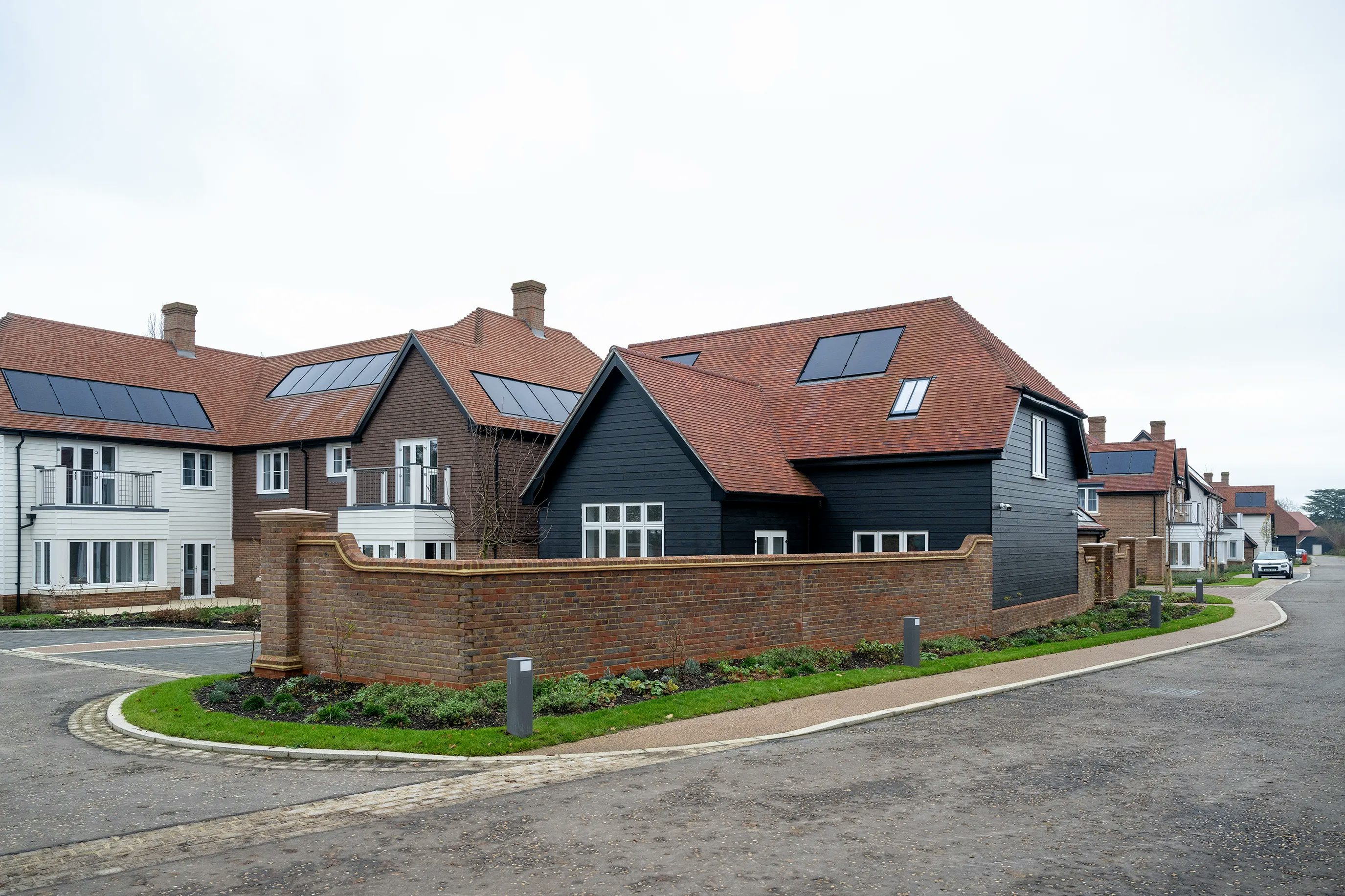 A modern residential street with black-clad, red-roofed houses—some featuring rooftop solar panels—lined along a gently curving, paved road. The foreground house, enclosed by a brick wall, has white-framed windows and a contemporary façade. Bollards and small patches of greenery border the pavement under a cloudy sky, creating a calm suburban atmosphere.