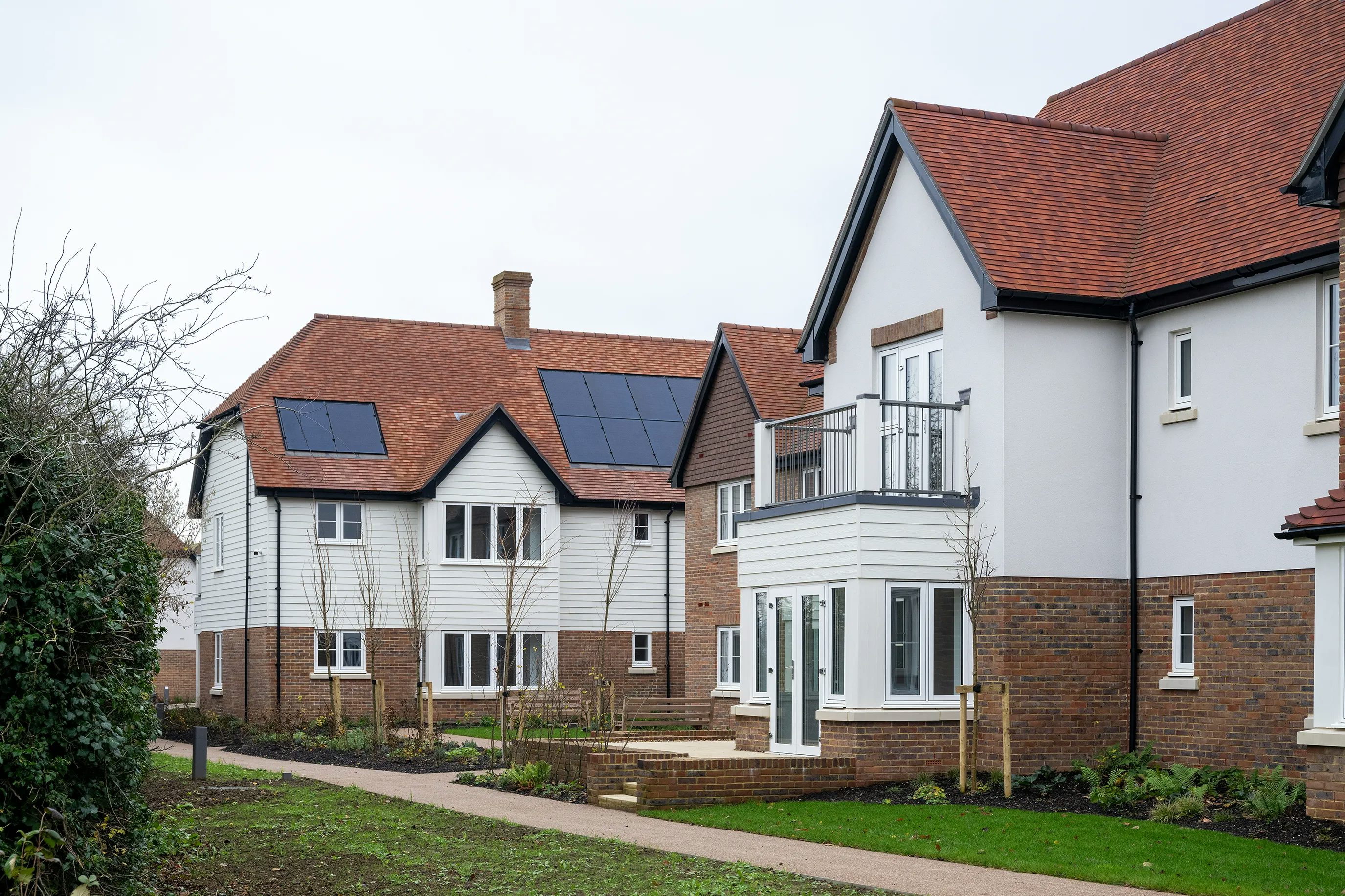 A row of two modern detached houses in a suburban setting, both with red-tiled roofs and white upper walls. The house on the left features rooftop solar panels, while the house on the right includes a small first-floor balcony with glass railings. Each home has a brick-clad ground floor, large windows, and landscaped front gardens with shrubs and young trees. A paved footpath runs in front, bordered by grass and framed by clear blue skies. The scene suggests an eco-conscious, family-friendly neighbourhood.