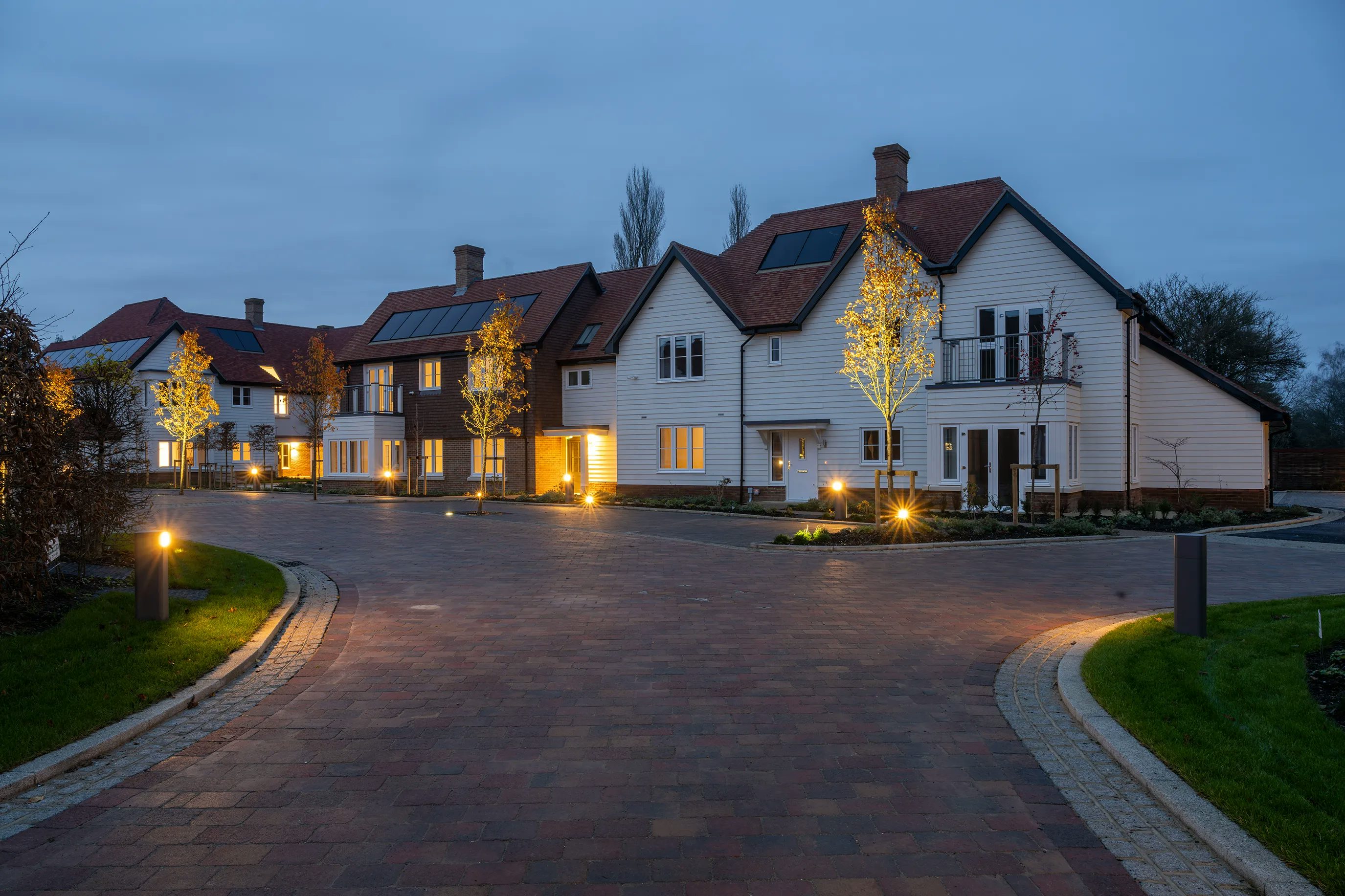 A dusk scene of a modern residential street featuring several detached houses with white and brown exteriors, pitched roofs fitted with solar panels, and warm ambient lighting. Trees line the paved driveways, subtly illuminated by street lamps and decorative lights, creating a serene, energy-efficient atmosphere that reflects sustainable suburban living. There are no visible people, vehicles, or movement, enhancing the calmness of the setting.