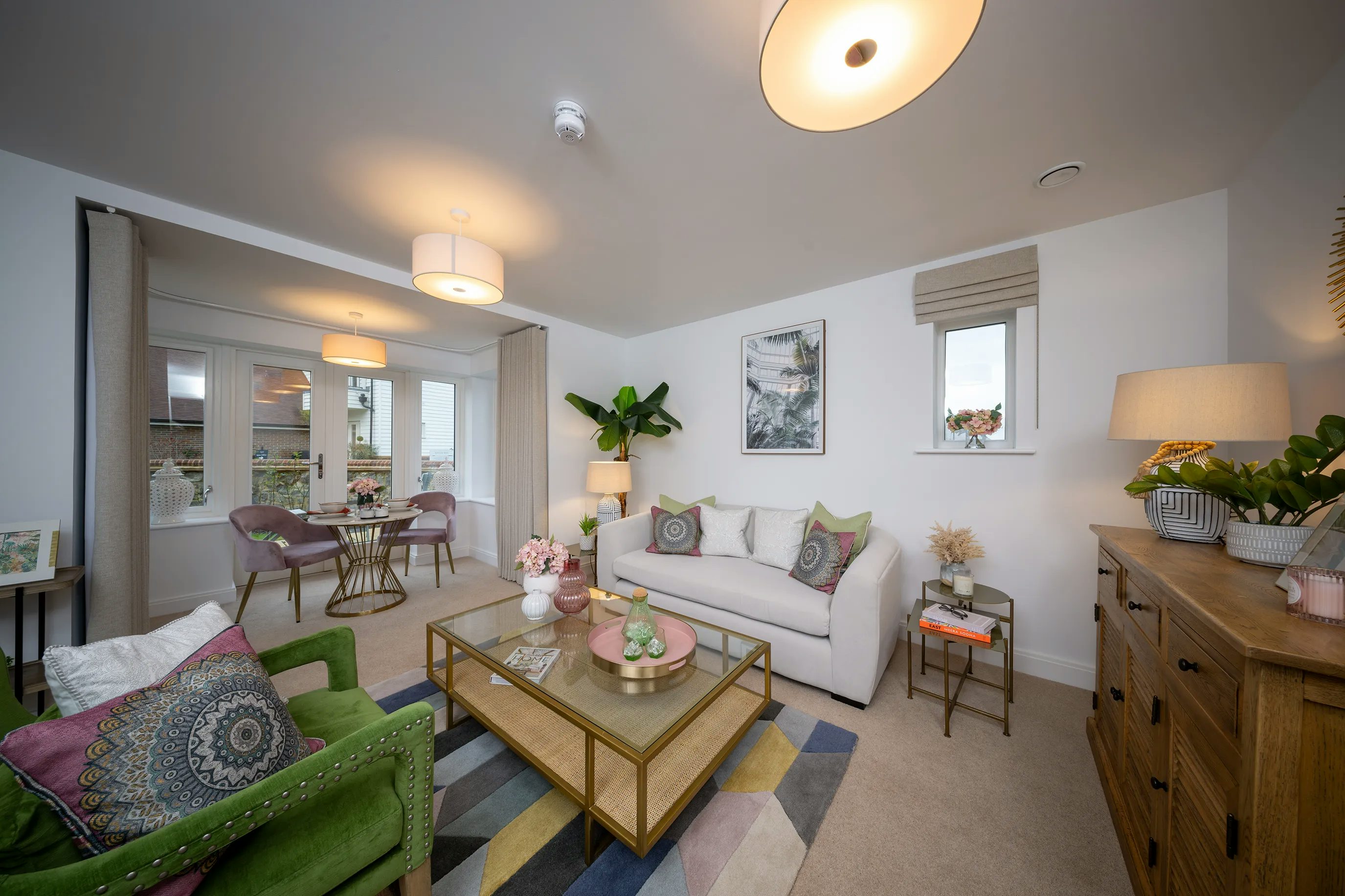 A contemporary open-plan living and dining room with a bright, inviting atmosphere. In the foreground, a white sofa with patterned cushions faces a wooden coffee table, adorned with decorative items. A green armchair adds contrast, positioned beside a wooden sideboard displaying houseplants and a lamp. A vibrant geometric rug anchors the seating area. To the rear, a circular dining table with pink upholstered chairs sits near large windows dressed with light curtains, allowing daylight to flood in. Framed artwork, modern lighting, and soft wood tones contribute to a stylish, comfortable interior.