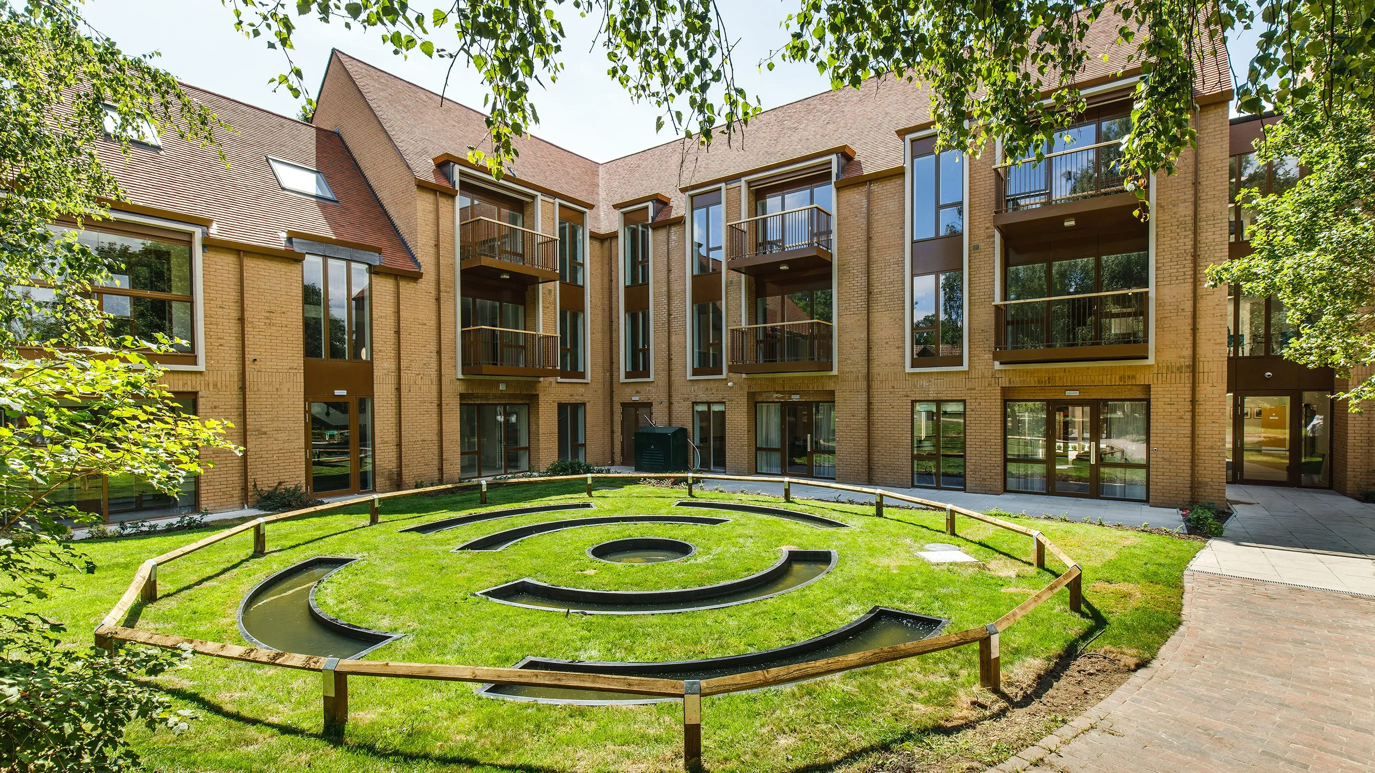 A three-storey modern residential building constructed with light brown brick, featuring large windows, private balconies, and a sloped roof with skylights. In the foreground, a circular landscaped garden with concentric stone paths, shallow water features, and trimmed greenery is enclosed by a low wooden fence. The surrounding lawn and trees create a peaceful, community-focused atmosphere.