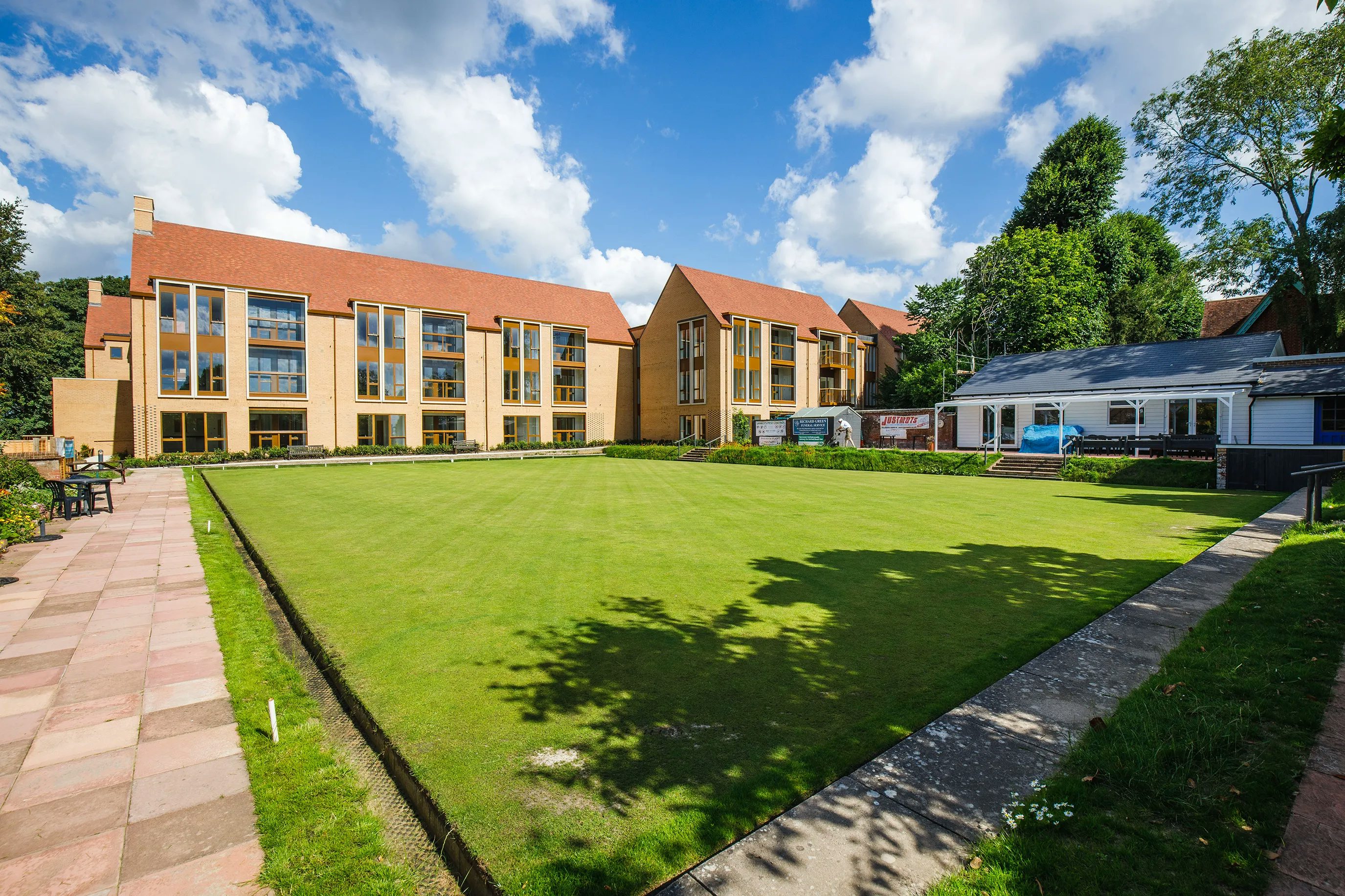 A manicured rectangular lawn bordered by paved walkways, framed by modern buildings with large windows and red roofs; a staircase leads to a smaller structure on the right, beneath a lightly clouded sky.