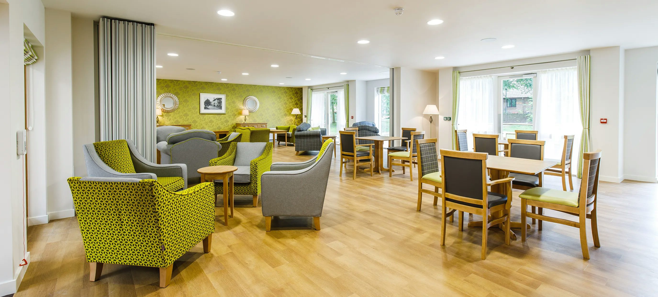 A spacious, sunlit communal room with wooden floors and large curtained windows. The space features clusters of wooden tables surrounded by green and grey upholstered chairs, arranged to encourage social interaction. Framed artwork, table lamps, and soft natural lighting create a warm, inviting atmosphere suggestive of a residential lounge or community centre.