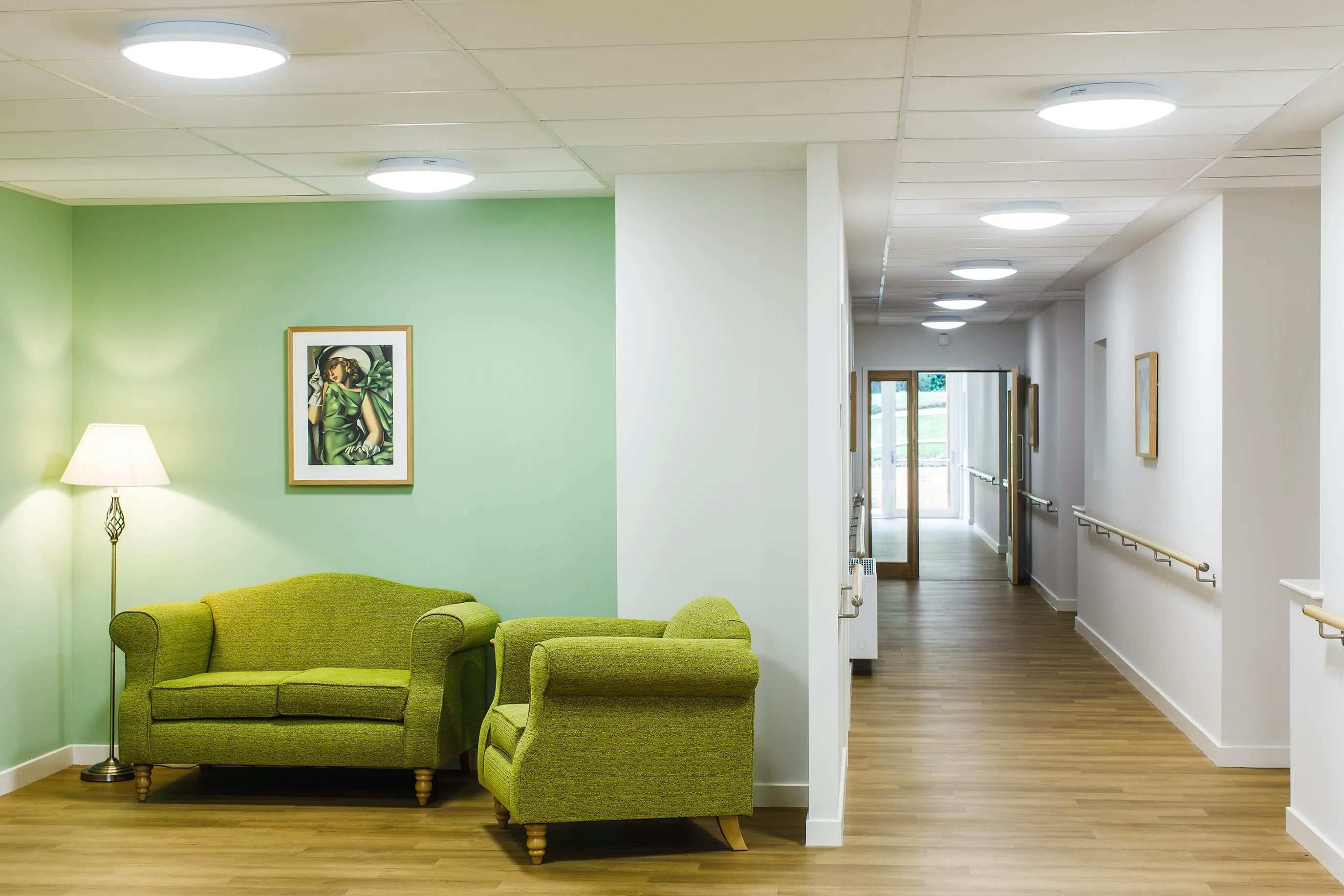 Bright, modern corridor in a healthcare or senior living facility featuring light wood flooring, soft green walls, and a cosy seating area with green upholstered chairs and a loveseat. A floor lamp and a framed portrait above the loveseat add warmth. The corridor extends into the distance with handrails on both sides and framed artwork along the walls, leading to glass doors at the far end, suggesting a safe, welcoming, and thoughtfully designed environment.