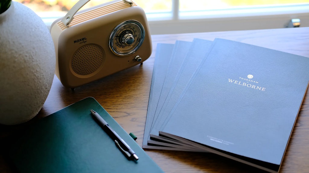 A wooden desk positioned in front of a window with greenery outside. On the left side sits a vintage-style ROXEL radio. To its right, four stacked blue booklets display “THAKEHAM WELBORNE” on the top one. In front of them lies a green notebook with a pen placed diagonally across it, suggesting a workspace or study area with a nostalgic, curated atmosphere.