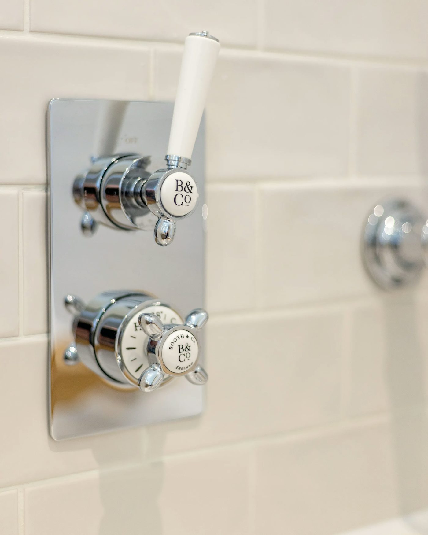 Close-up of a chrome B&CO shower control panel mounted on a tiled bathroom wall, featuring two knobs—one above with a smooth white cylindrical handle, and one below with a cross-shaped metallic design and a visible temperature indicator.