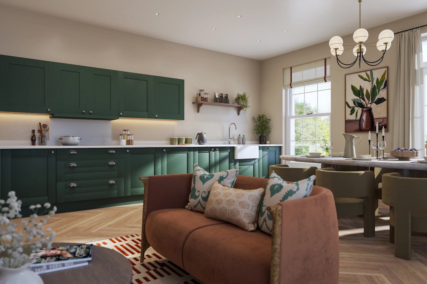 A warm, modern kitchen and dining space with green cabinetry, white countertops, and silver accents. A wooden dining table sits beneath a chandelier beside a large window, flooding the room with natural light. The kitchen features open shelving with plants and decorative items, while the foreground includes a cosy seating area with a brown sofa, patterned cushions, and a coffee table topped with a vase of fresh flowers and a magazine. The overall atmosphere is inviting, blending rustic charm with contemporary design.