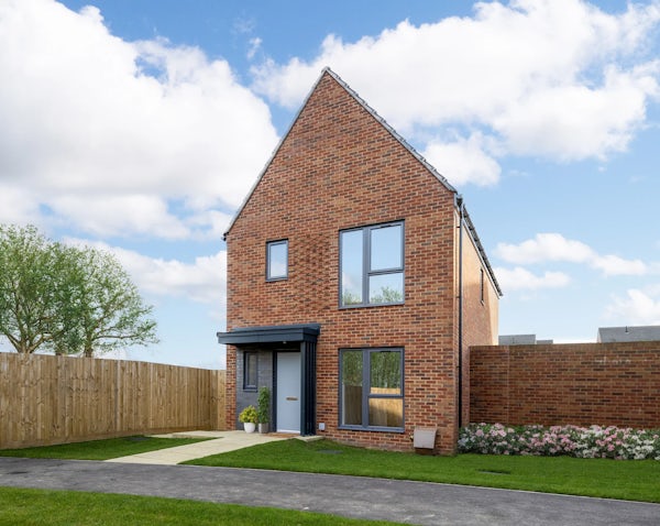 A two-storey modern brick house with a steep triangular roof and large front-facing windows. A black front door is flanked by a small potted plant, with a wooden fence on the left and a brick boundary wall on the right. The front garden features neat green grass, a paved path, and a flower bed along the wall. A leafy tree rises behind the fence, under a blue sky with light clouds.