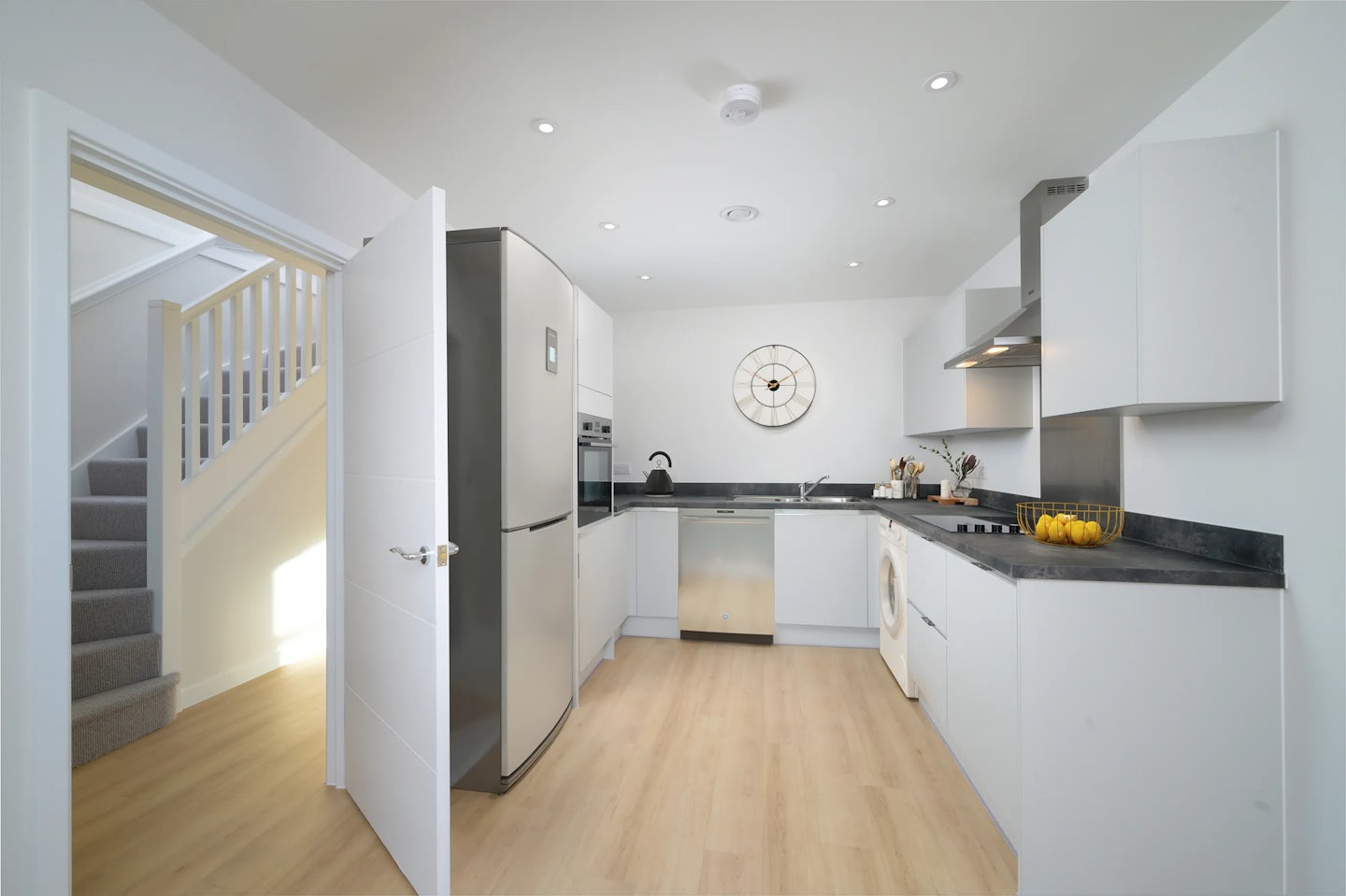 A modern kitchen interior featuring sleek white cabinets and integrated stainless steel appliances, including a refrigerator, oven, dishwasher, washing machine, and a stovetop with an overhead extractor. A decorative wall clock hangs above the sink, while warm wood flooring and recessed ceiling lights contribute to a clean, well-lit atmosphere. To the left, an open door reveals a carpeted staircase leading upward, suggesting a residential setting.