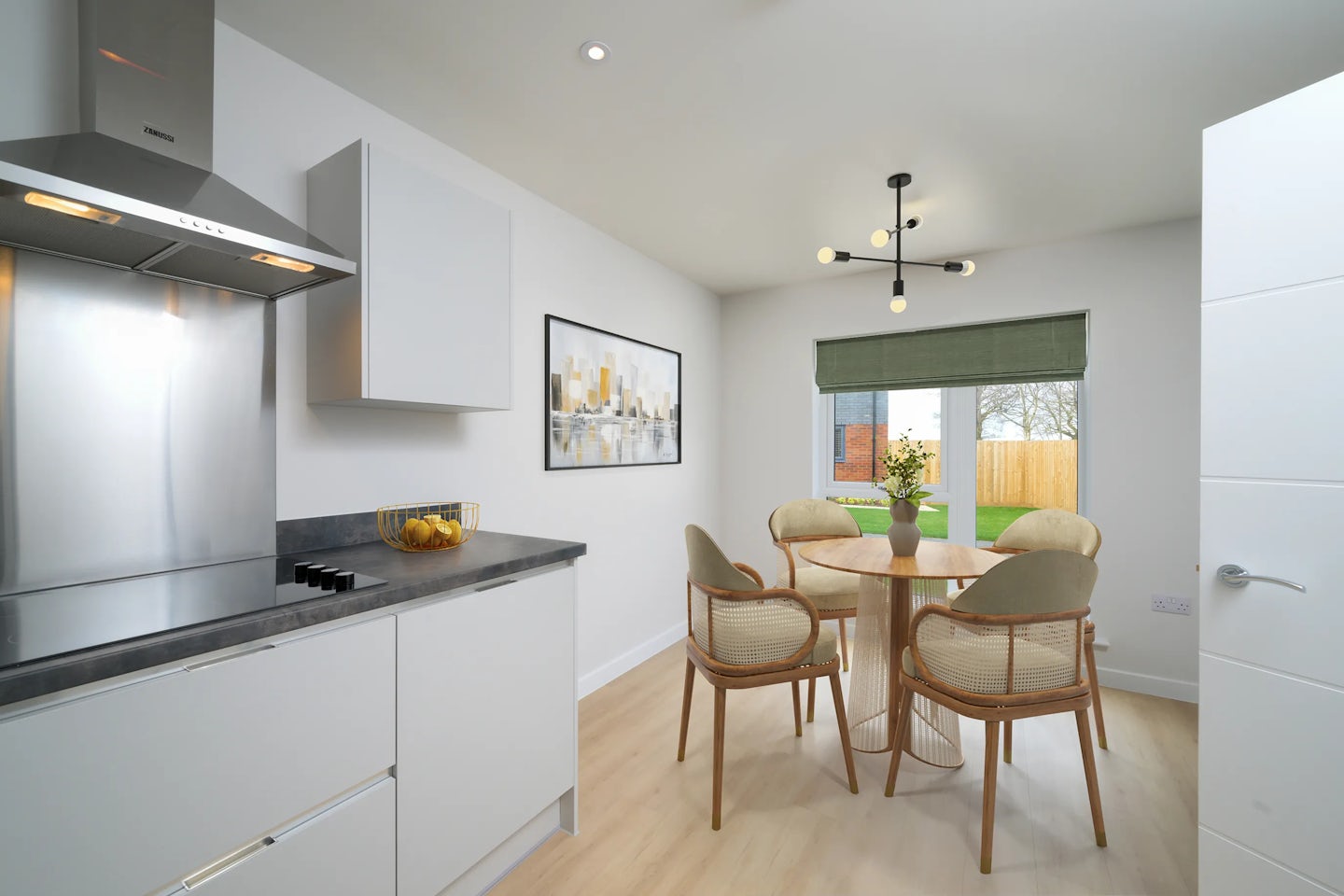 A bright, contemporary kitchen and dining area. The kitchen features white cabinetry, a black countertop with a bowl of lemons, and a stainless steel range hood. The dining space includes a round wooden table set with a vase of flowers and four wicker chairs. A modern light fixture hangs above the table. Large windows with green blinds reveal a garden view with trees and a wooden fence. A framed artwork is mounted on the wall between the kitchen and dining zones.