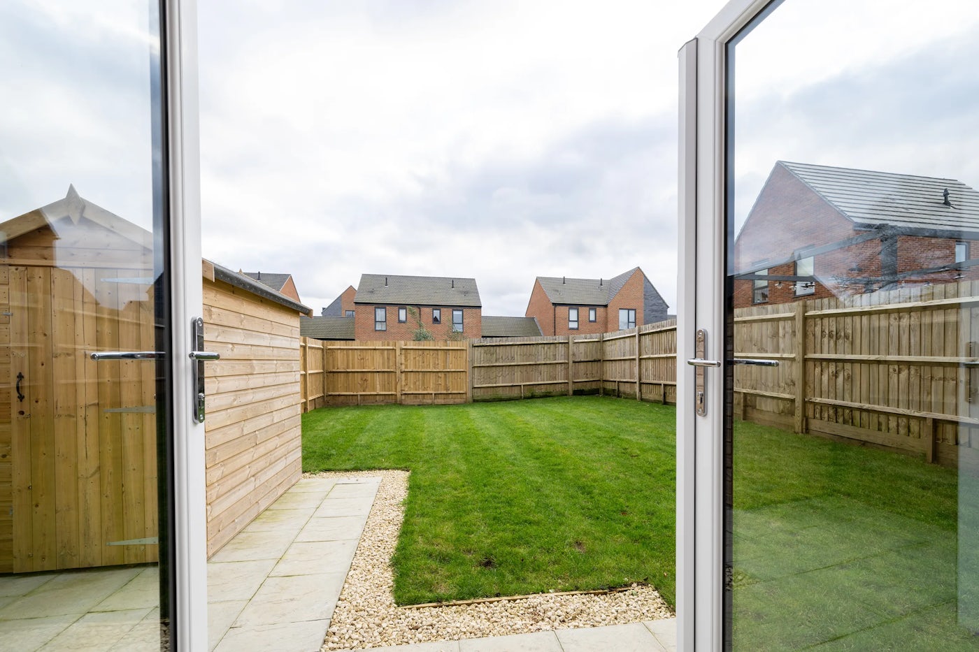 A view of a suburban back garden seen through open double glass doors. The garden features a lush green lawn enclosed by a wooden fence, with a wooden shed to the left. Beyond the fence, modern brick houses with pitched roofs line the background under a cloudy sky, creating a peaceful, overcast suburban atmosphere.