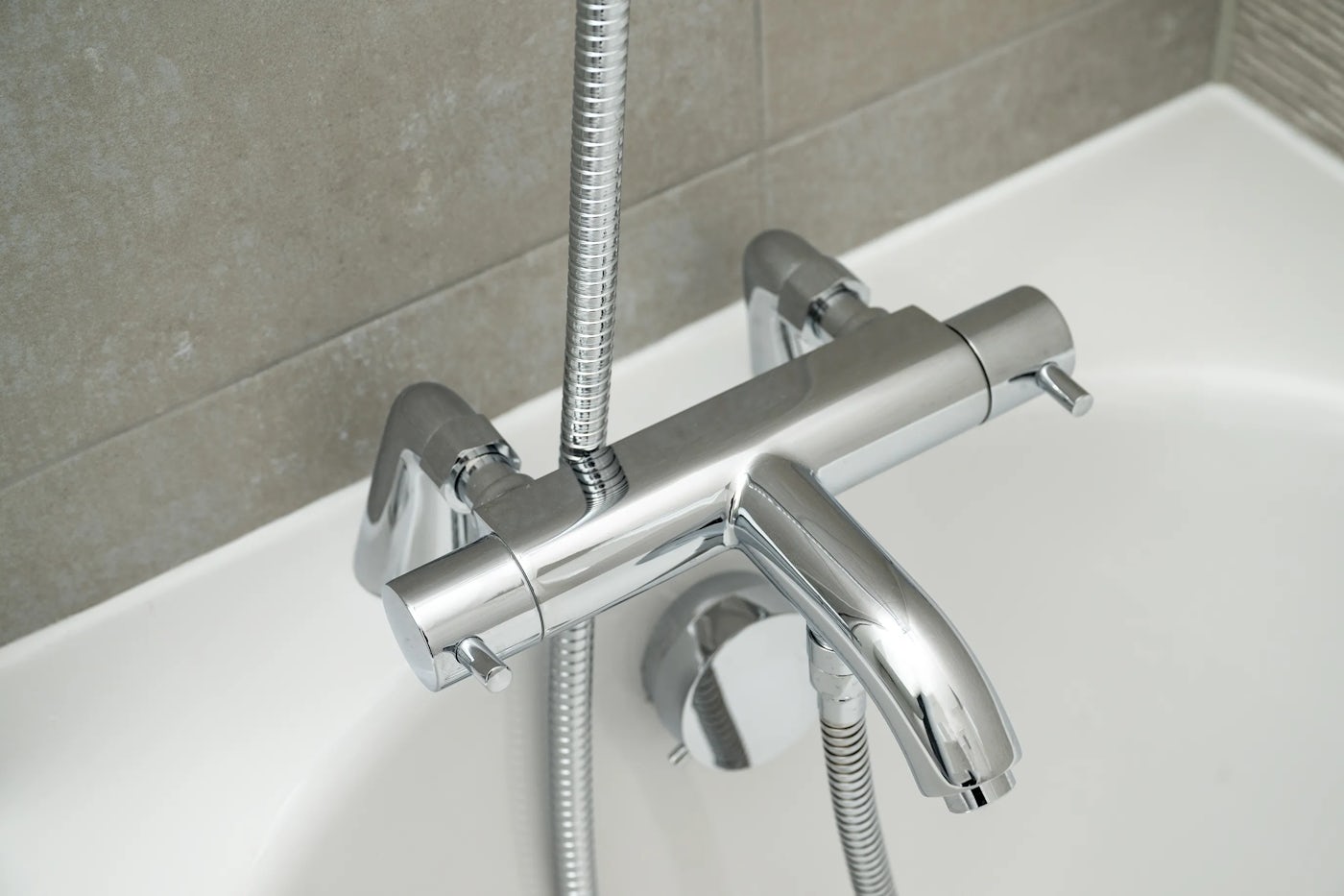 Close-up of a chrome-finished bathtub faucet with a lever handle and an attached handheld showerhead, positioned against a tiled bathroom wall. The showerhead rests at the edge of the tub, its flexible hose looped neatly below the fixture, reflecting a minimalist and modern aesthetic.