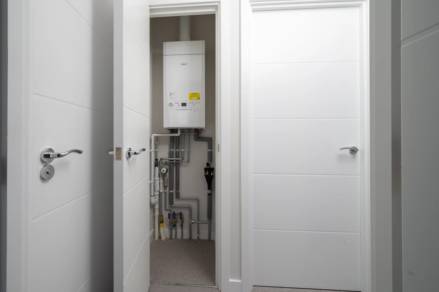 A compact utility room with white walls and door frames, featuring an open cupboard that reveals a wall-mounted boiler system. The boiler has visible copper and white piping, valves, and gauges neatly connected beneath it. The space appears clean and well-maintained, with a simple, functional design.