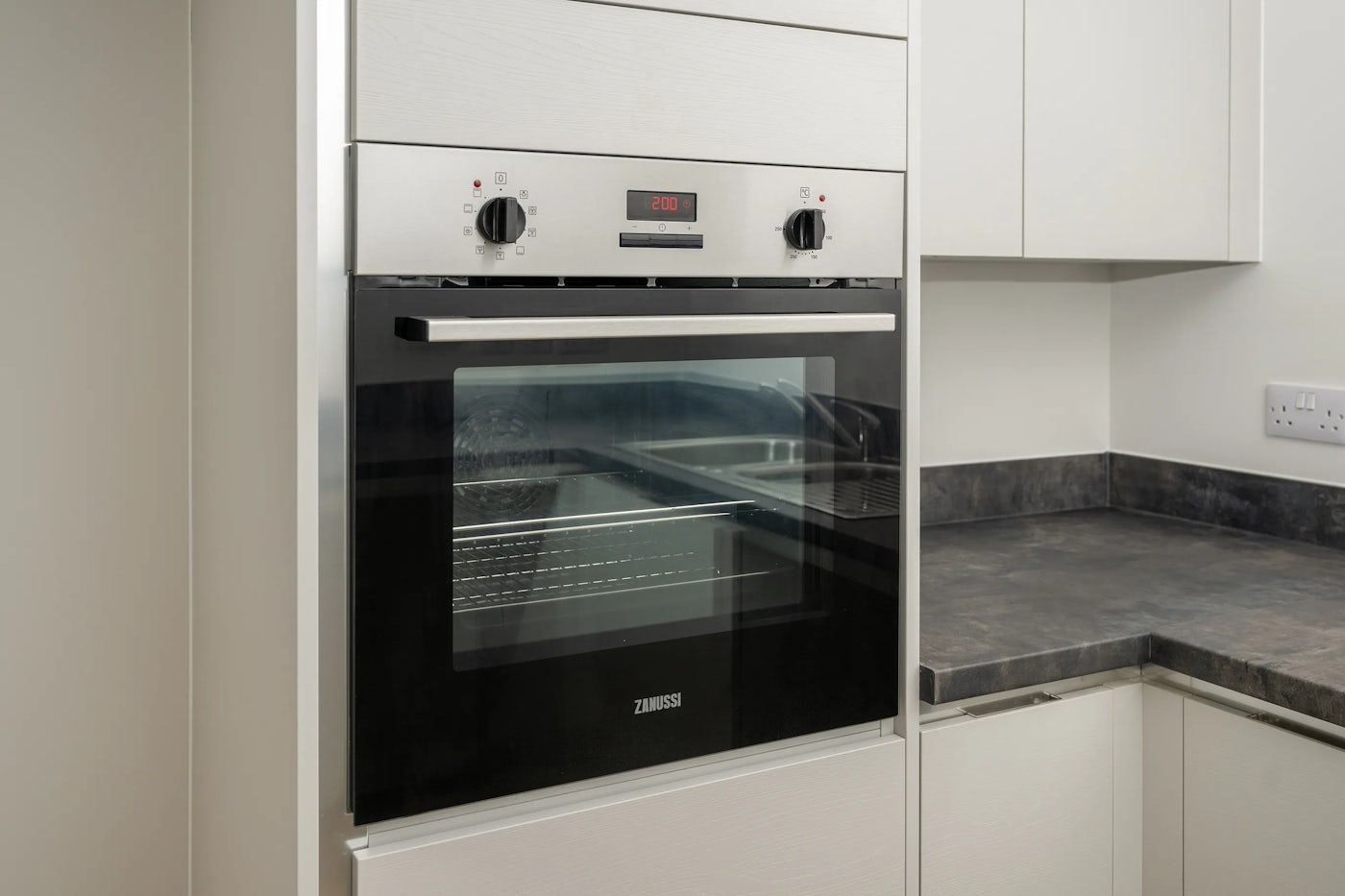 A built-in Zanussi oven with a black glass door and digital display set to "20:00," flanked by two silver control knobs. The oven is integrated into sleek white kitchen cabinetry with a dark countertop above, reflecting a modern and minimalist design