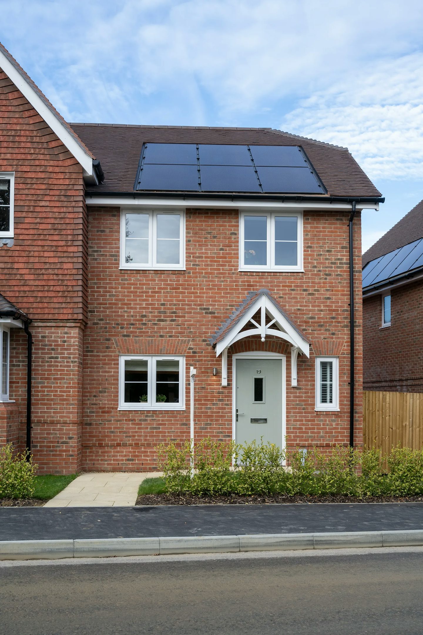 Modern two-storey brick house with solar panels on the pitched roof, a white front door sheltered by a timber-framed overhang, and four white-framed windows. Similar homes are visible in the suburban background, suggesting a planned residential development focused on sustainability.