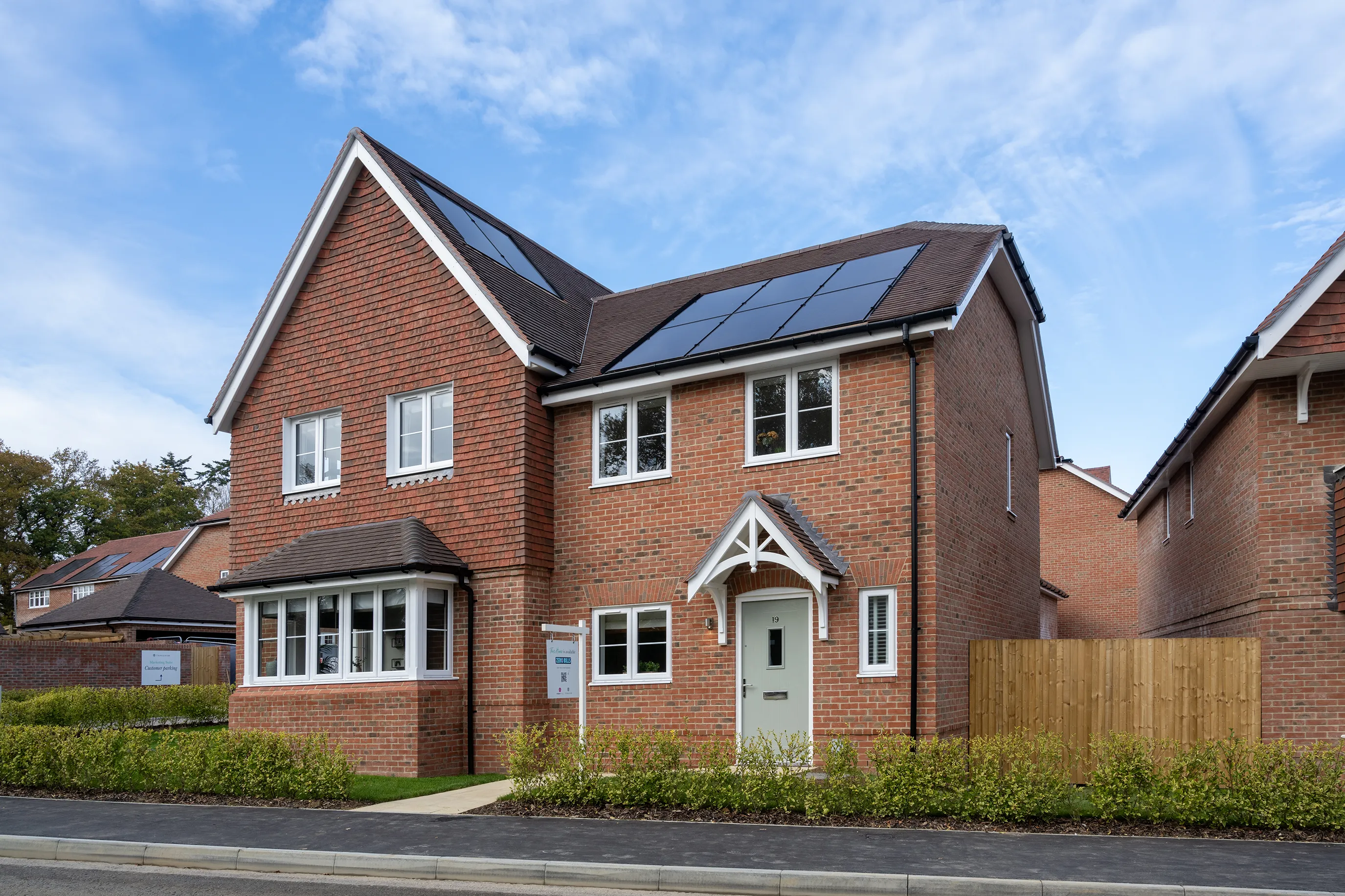 A modern two-storey brick semi detached house with solar panels on the roof, a white-framed front door with a small canopy, and multiple windows including a bay window on the left. A neatly trimmed hedge runs along the front, framing a paved path to the entrance. The scene is set under a partly cloudy sky, with similar houses visible in the background, suggesting a well-maintained residential area focused on sustainability.