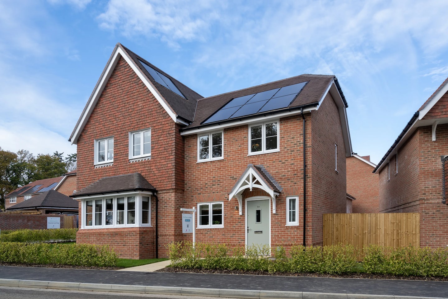 A modern two-storey brick semi detached house with solar panels on the roof, a white-framed front door with a small canopy, and multiple windows including a bay window on the left. A neatly trimmed hedge runs along the front, framing a paved path to the entrance. The scene is set under a partly cloudy sky, with similar houses visible in the background, suggesting a well-maintained residential area focused on sustainability.