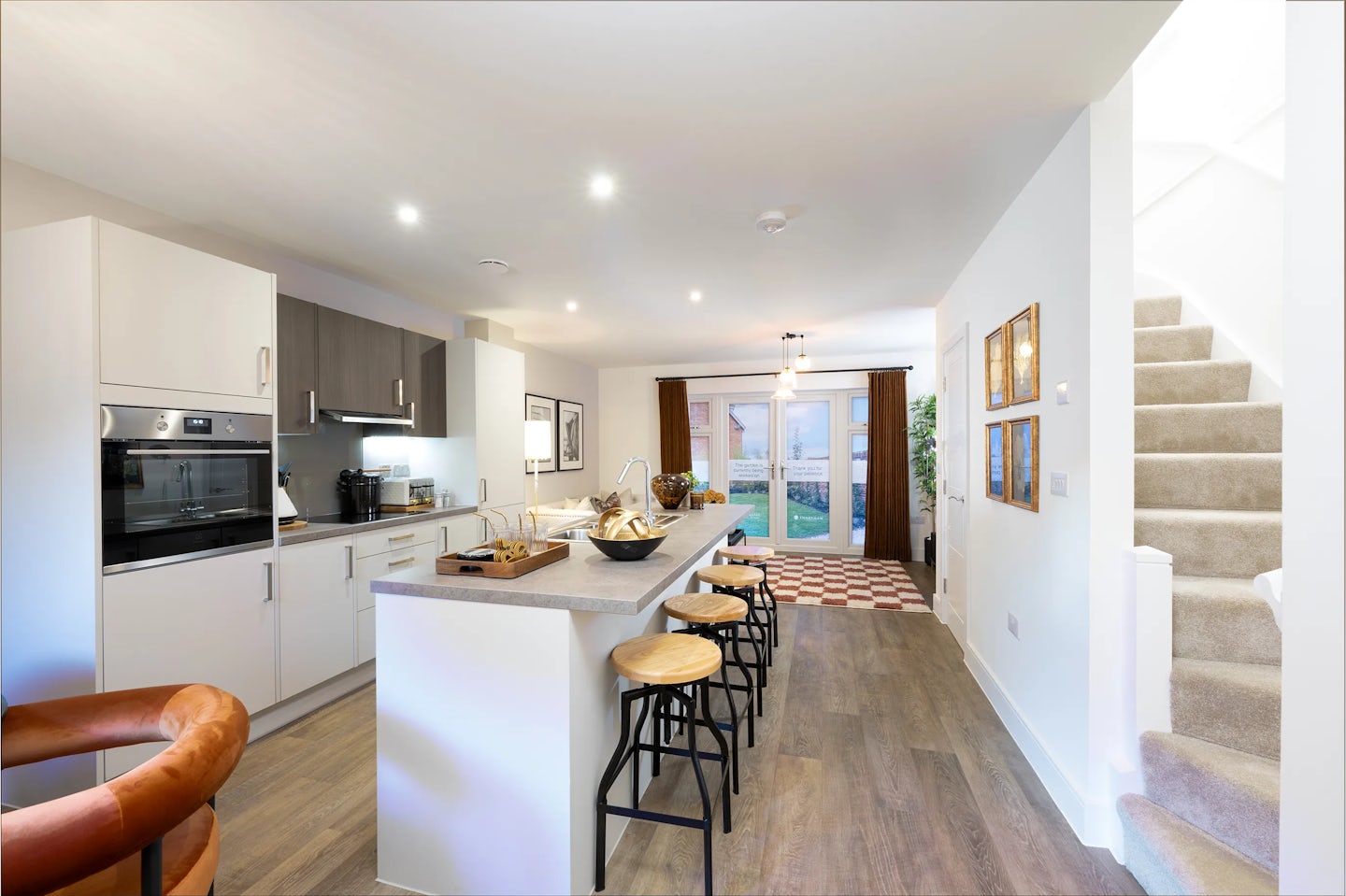 A bright, modern kitchen with white cabinetry, a built-in oven, and a central island featuring a light countertop and bar stools. Bowls, a cutting board, and other items are arranged neatly atop the island. In the background, a dining table and chairs sit near large windows that allow natural light to fill the space. To the right, a staircase leads to an upper level.