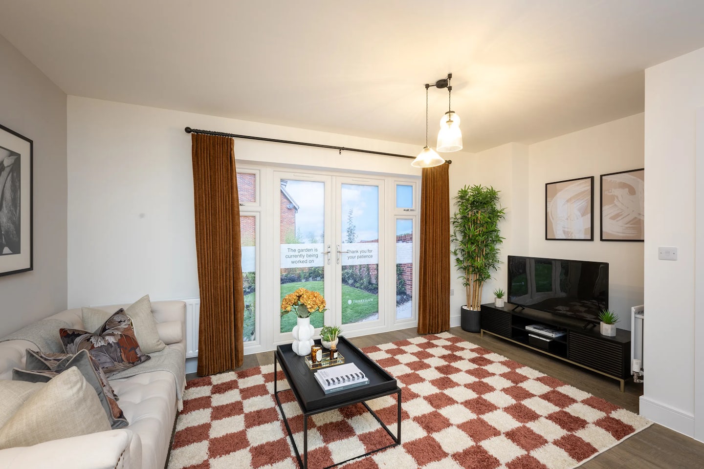 A modern living room with a relaxed, welcoming atmosphere. On the left, a white sofa is adorned with patterned and textured cushions. In the centre, a rectangular wooden coffee table displays a tray, stacked books, and a small plant. To the right, a television sits on a sleek black stand beside a tall leafy houseplant. The room is softly lit by two pendant lights and natural daylight streaming through a large window with brown curtains, overlooking a green garden. A checkered red-and-white rug covers the wooden floor.