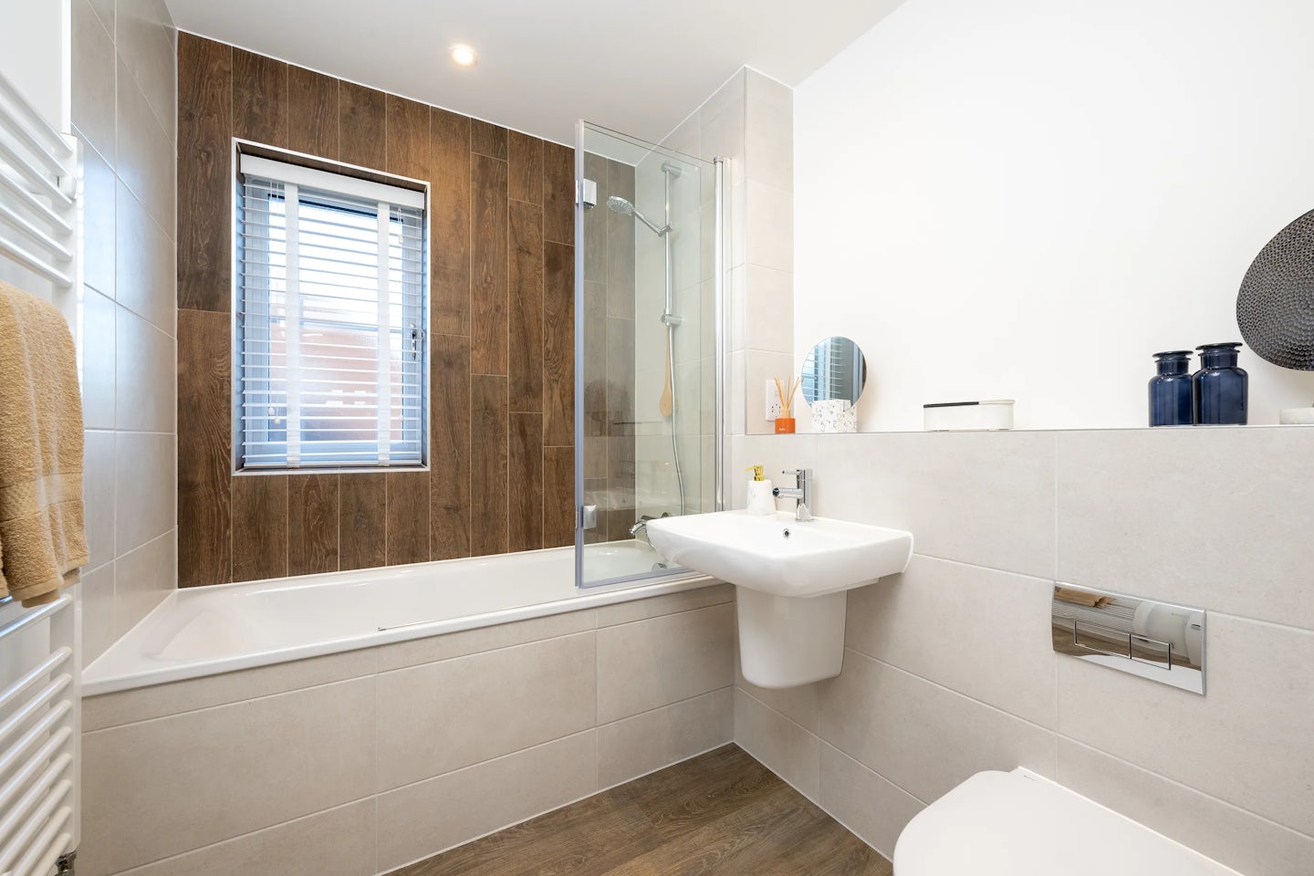 A contemporary bathroom featuring a bathtub with a glass shower screen on the left, backed by wood-effect wall tiles and a window with blinds. A wall-mounted sink with a mirror and shelf of toiletries sits beside the tub, while a partially visible toilet is on the right. The space includes a wood-style floor, a towel draped over a radiator, and overhead lighting, creating a clean, modern atmosphere.