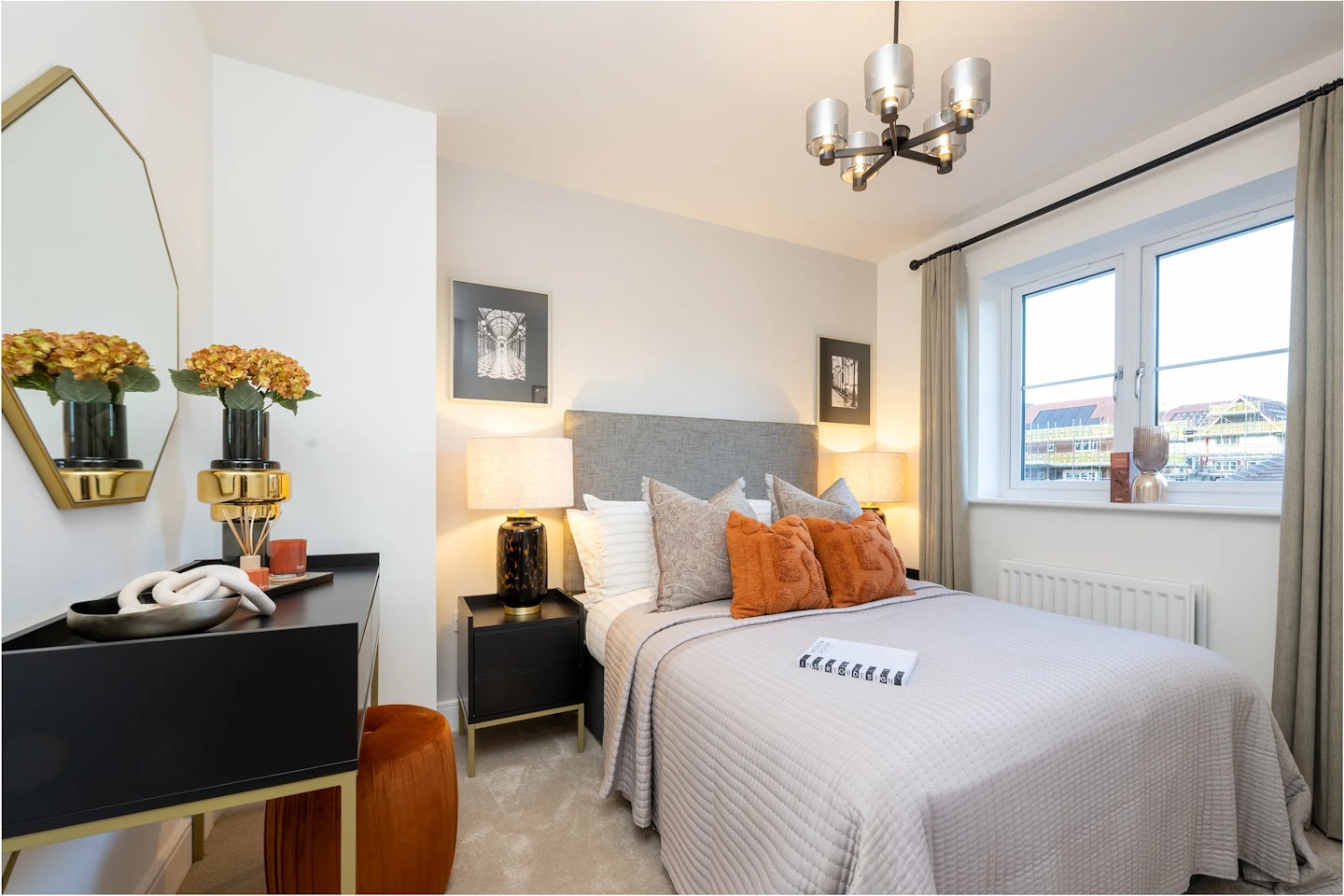 Modern bedroom with a soft, neutral colour palette. A double bed with a light grey quilted duvet and layered pillows, including orange and charcoal, sits centrally against a wall with two minimalist framed prints above. On either side, matching bedside tables each hold a lamp and small decorative items. A closed notebook lies on the bed. To the left, a black vanity table features a round mirror, floral arrangements in ceramic vases, and assorted accessories, paired with a small orange stool beneath. A large window with beige curtains brings in natural light, and a contemporary chandelier hangs from the ceiling, adding an elegant touch.