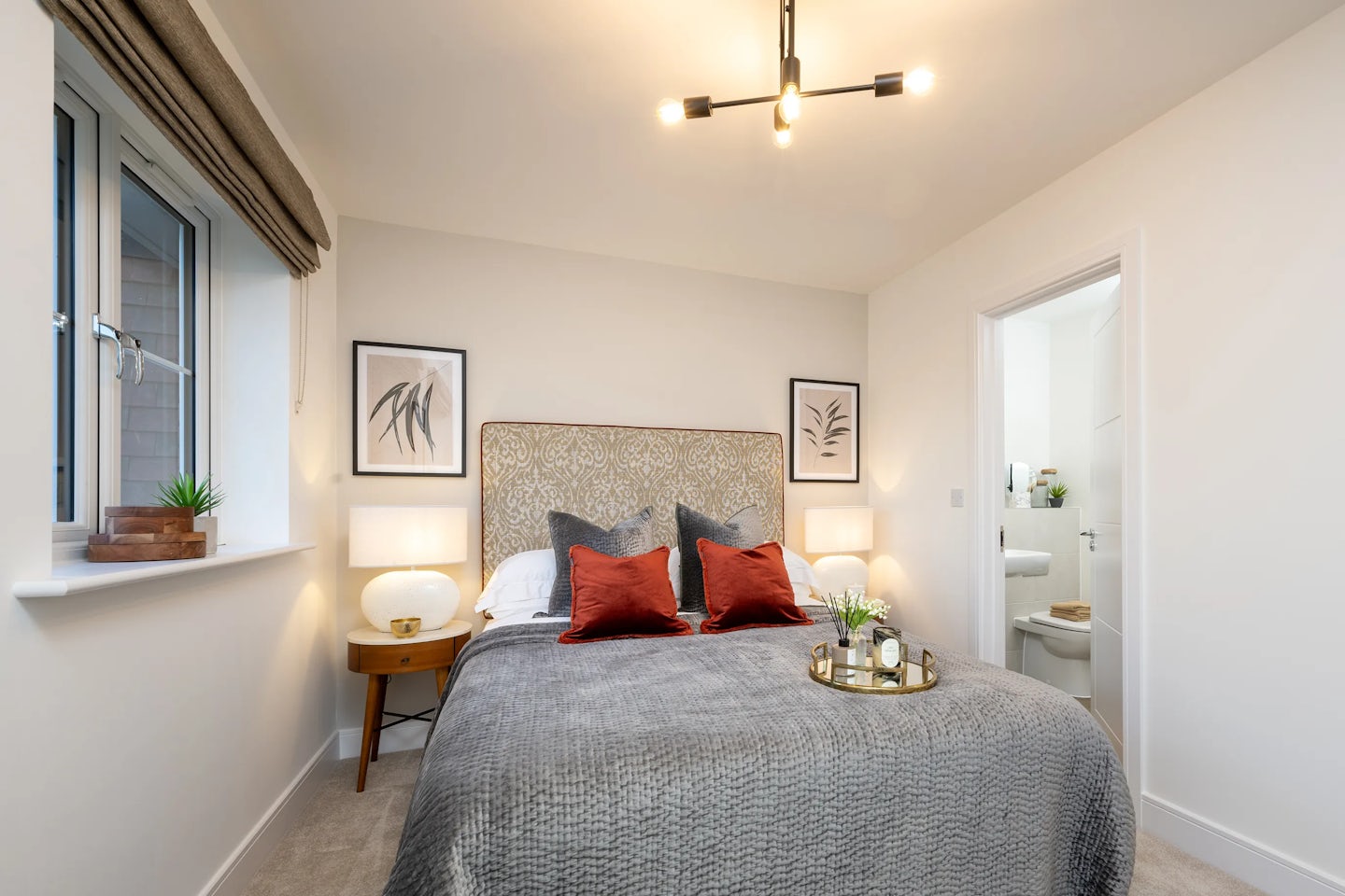 Modern bedroom featuring a centrally placed double bed with a grey textured blanket and a mix of red and grey pillows. Two matching bedside tables hold contemporary lamps and small decorative objects. Above the bed hang two framed botanical prints. A window with a brown Roman shade is on the left wall, while a door on the right leads to an en-suite bathroom. A sculptural ceiling light fixture adds a stylish focal point to the minimalist, cosy atmosphere.