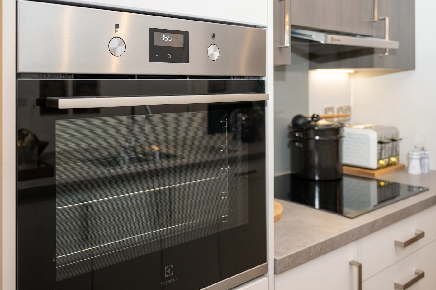 A modern kitchen interior with a stainless steel built-in oven set within light cabinetry. The oven displays the time "15:55" on a digital screen and features two round control knobs.