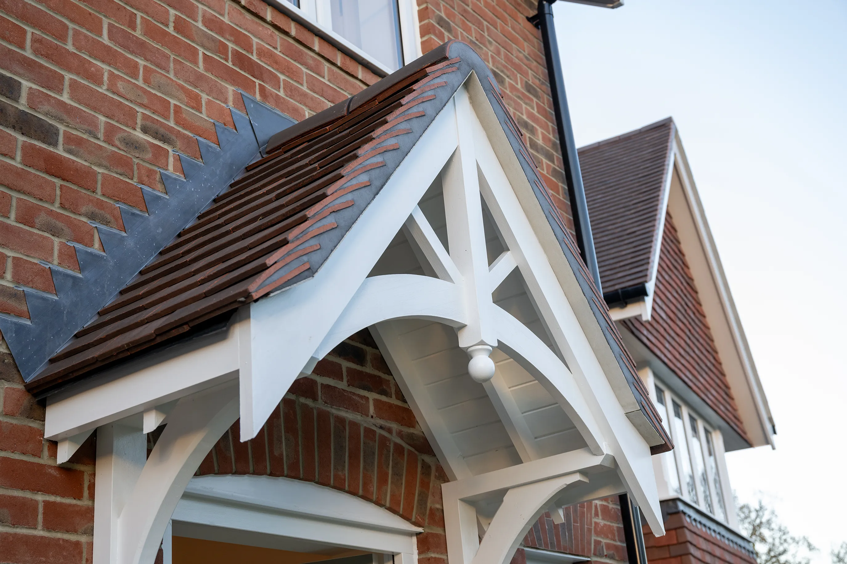 Front elevation of a red-brick house showing a gabled porch roof with white timber trim, brown roof tiles, and a decorative circular ornament at the gable peak. A window is visible above the porch.