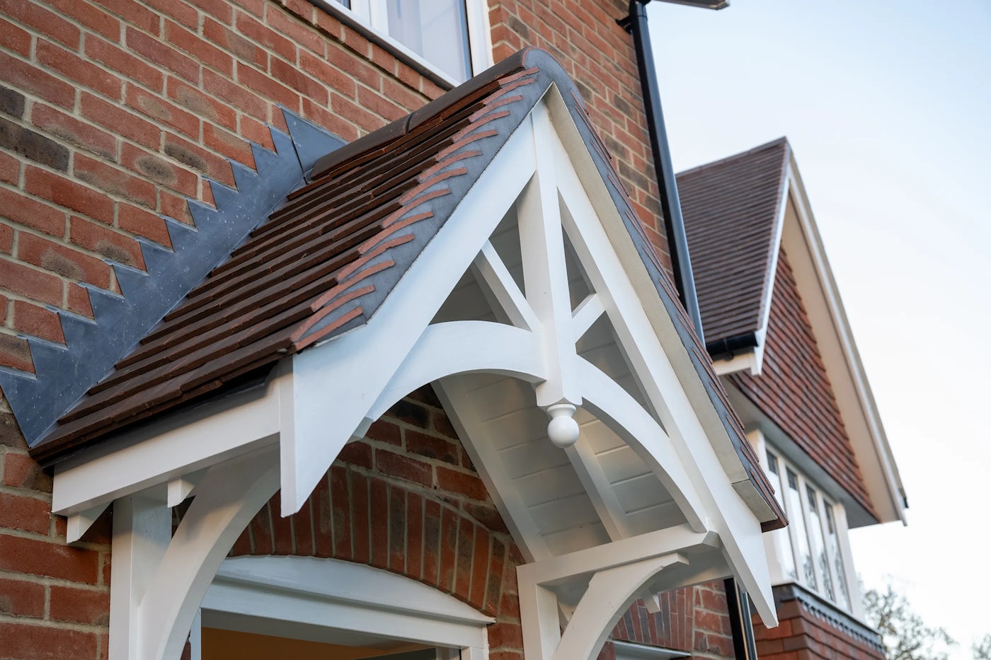 Front elevation of a red-brick house showing a gabled porch roof with white timber trim, brown roof tiles, and a decorative circular ornament at the gable peak. A window is visible above the porch.