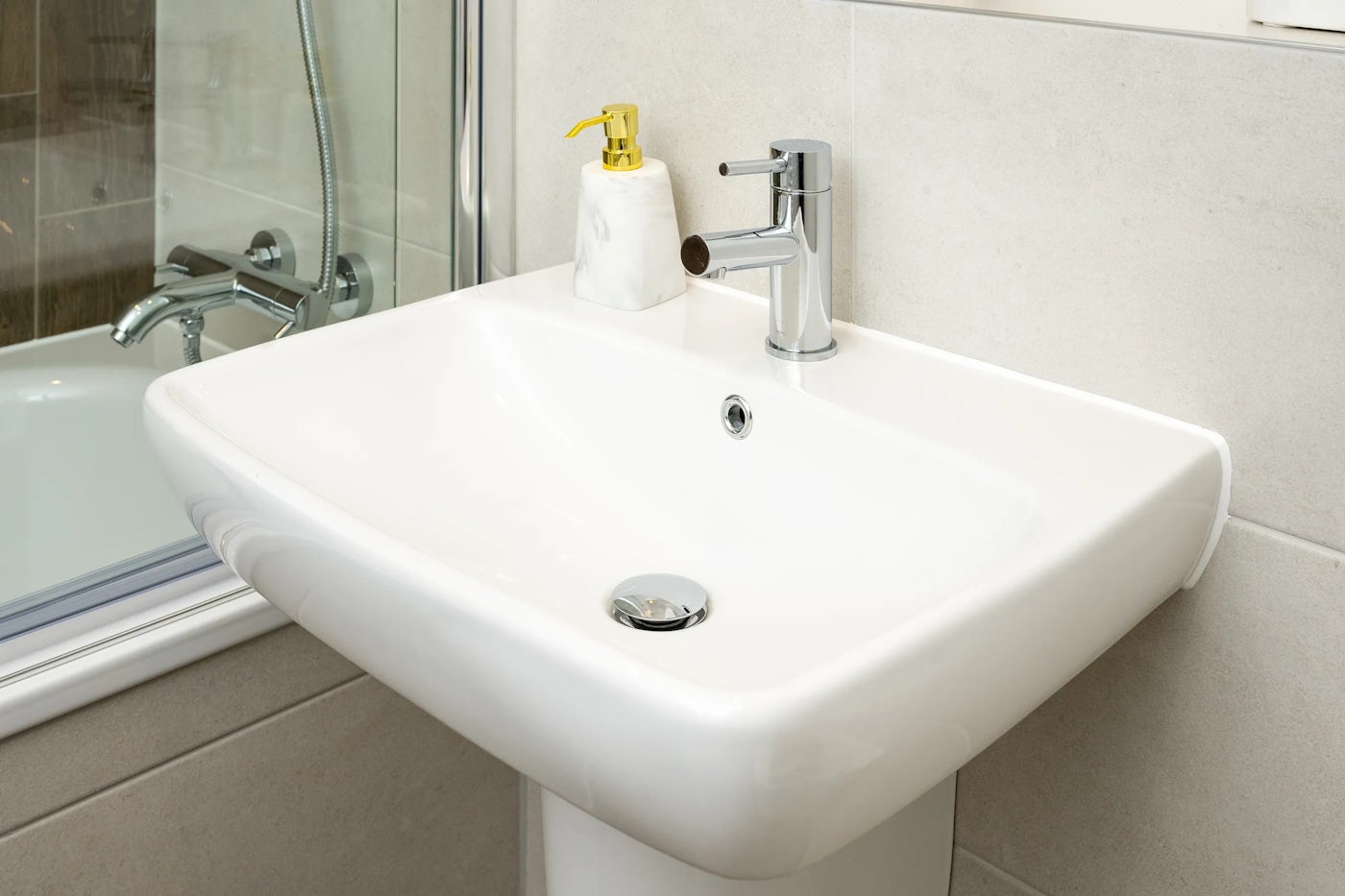 Modern bathroom sink with a rectangular white basin and chrome faucet, paired with a marble-style soap dispenser featuring a gold pump. A glass partition reveals a bathtub behind the sink, with a chrome showerhead and faucet contributing to the clean, contemporary design.