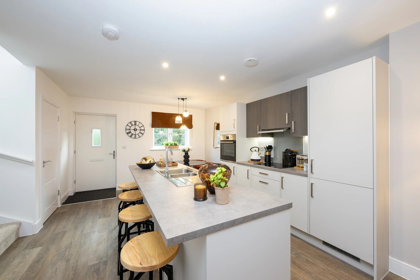 A contemporary kitchen featuring a central island with a built-in sink and four wooden stools with black metal legs. The island is styled with a small plant, candle, and bowl of fruit, adding a warm, lived-in touch. White cabinetry with stainless steel handles lines the back wall, housing a built-in oven, microwave, and coffee machine. A window above the sink is fitted with a brown blind, and a round clock hangs near a white entrance door. Wood-effect flooring and a mix of recessed and pendant lighting contribute to the room’s bright, welcoming feel.