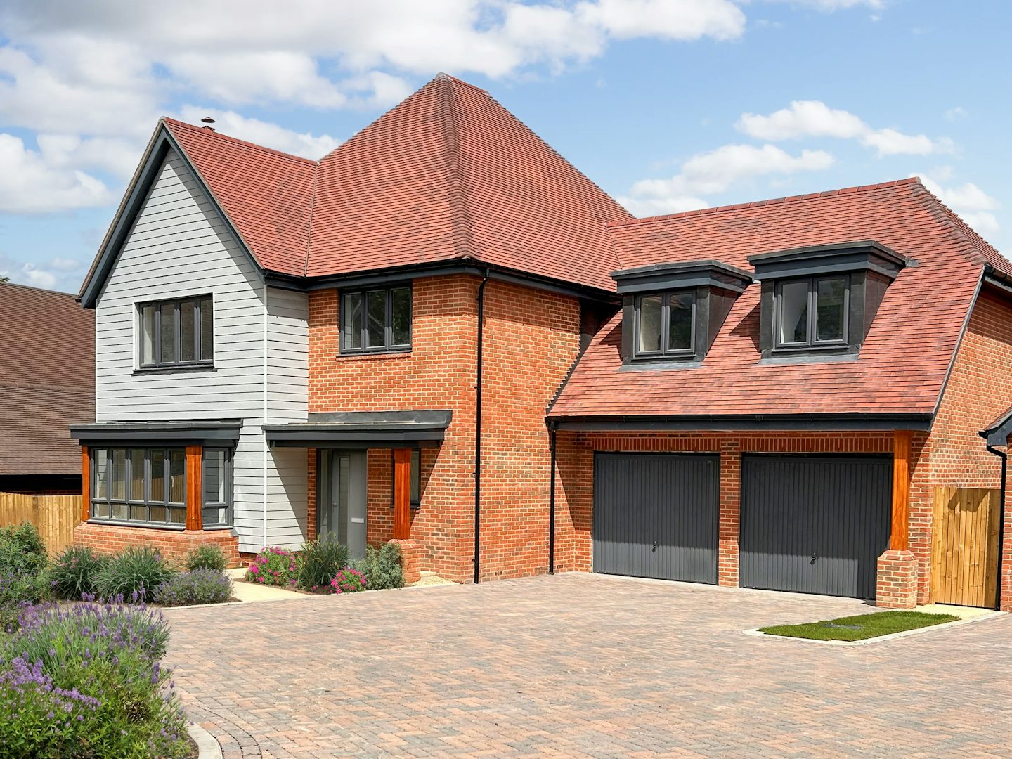 A modern two-storey suburban house with a red-tiled roof, brick and grey siding exterior, and two dormer windows above a double garage. The paved brick driveway leads to landscaped front gardens with flowering plants, set beneath a partly cloudy sky.