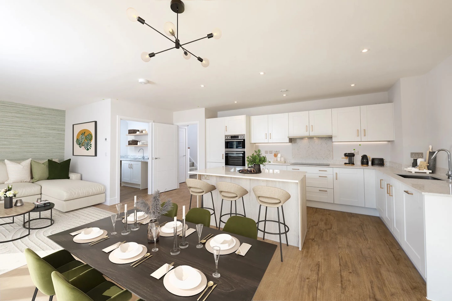 A spacious, modern open-plan kitchen and dining area with a minimalist aesthetic. The kitchen features sleek white cabinetry and a large central island with four wooden bar stools. Appliances such as a kettle, toaster, and built-in oven are neatly arranged. Adjacent to the kitchen, a dark wood dining table is set for a meal, surrounded by green velvet chairs. In the background, a cosy living space includes a white sofa, a low coffee table, and wall-mounted artwork. The room is brightened by natural daylight and ceiling spotlights, with light wood flooring unifying the open layout.