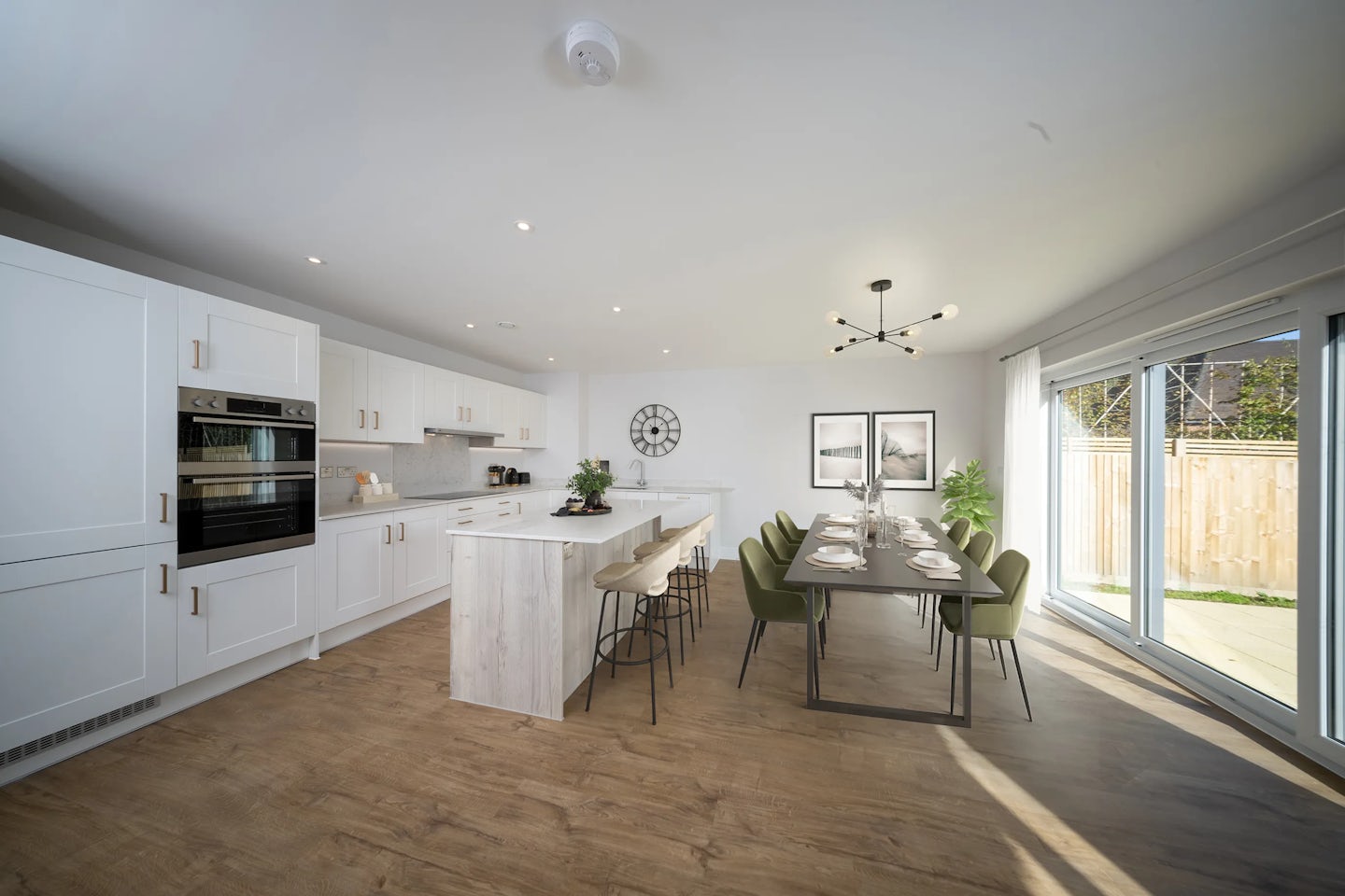A modern open-plan kitchen and dining area featuring white cabinetry, a central island with bar stools, and a built-in oven. The dining space includes a long table set for a meal, surrounded by green chairs. Large windows on the right flood the room with natural light, while decorative plants and framed wall art add warmth. Light wood flooring and a clean, contemporary design evoke a welcoming, airy atmosphere.